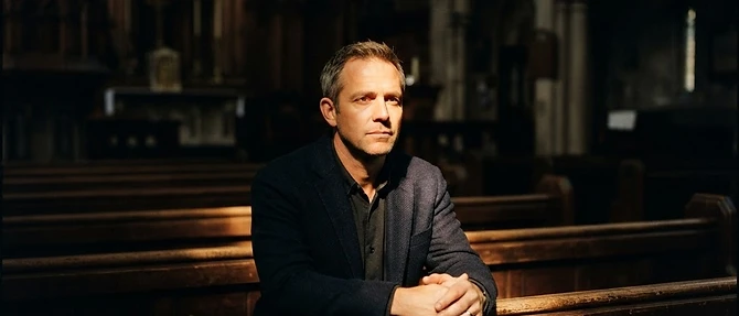 A man sitting alone in a dark church pew with one shaft of warm light falling across him