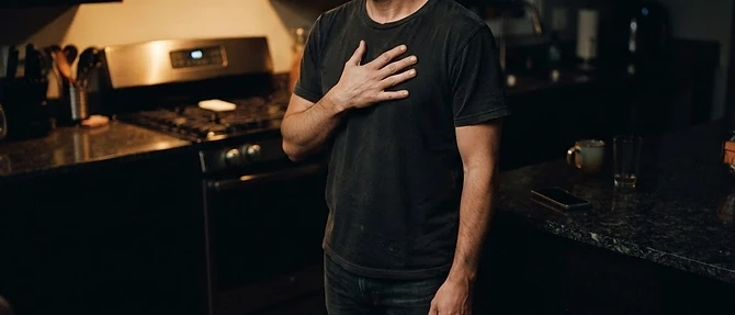 A man standing still in a dim kitchen at night, hand pressed to his chest, breathing slowly