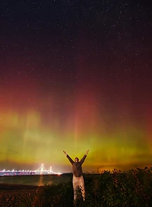 Person with arms outstretched under an aurora beside the lit Mackinac Bridge