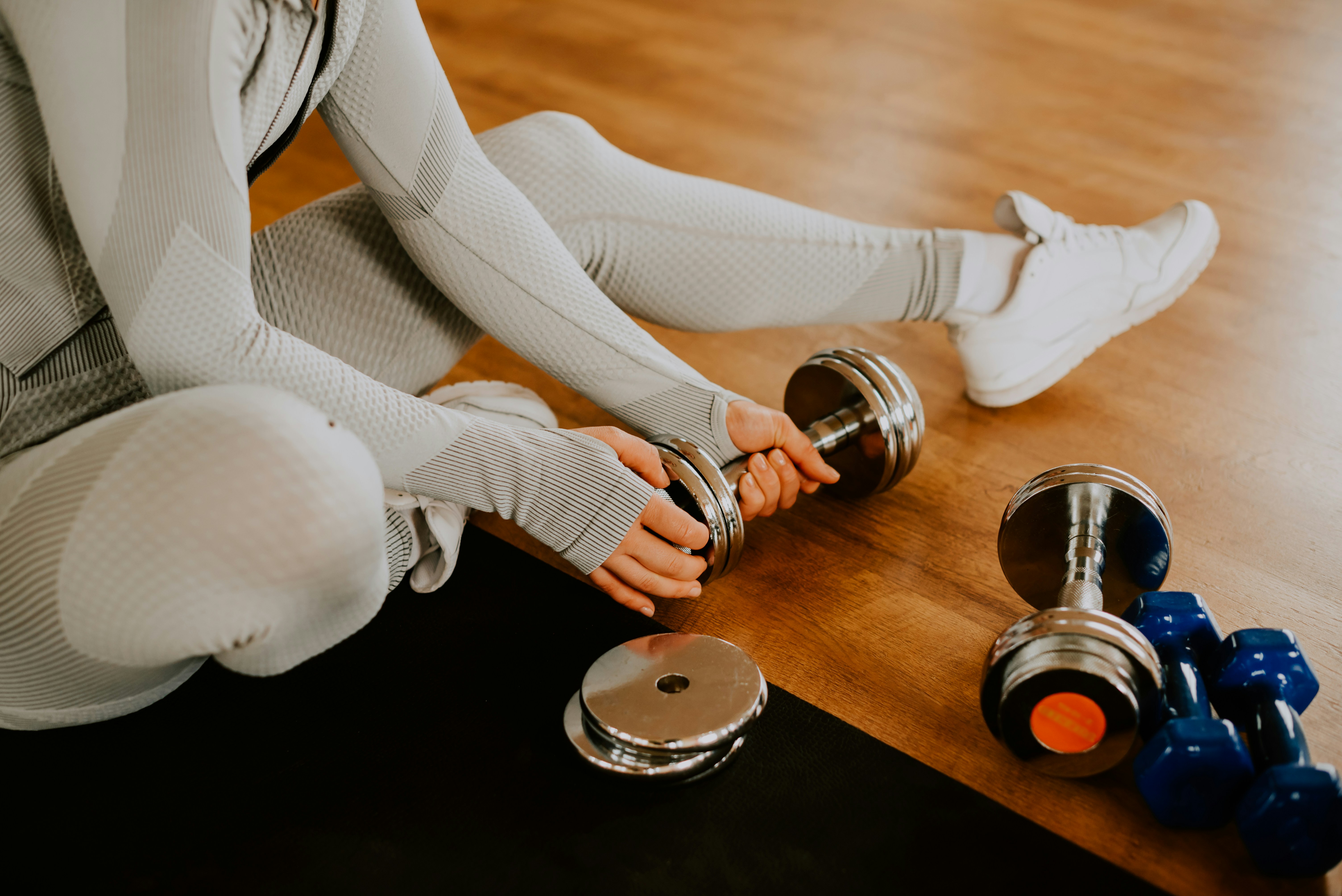 A person seated on a gym floor in light grey athletic wear holding a chrome dumbbell with both hands, with blue and chrome dumbbells on a warm wood laminate floor beside them