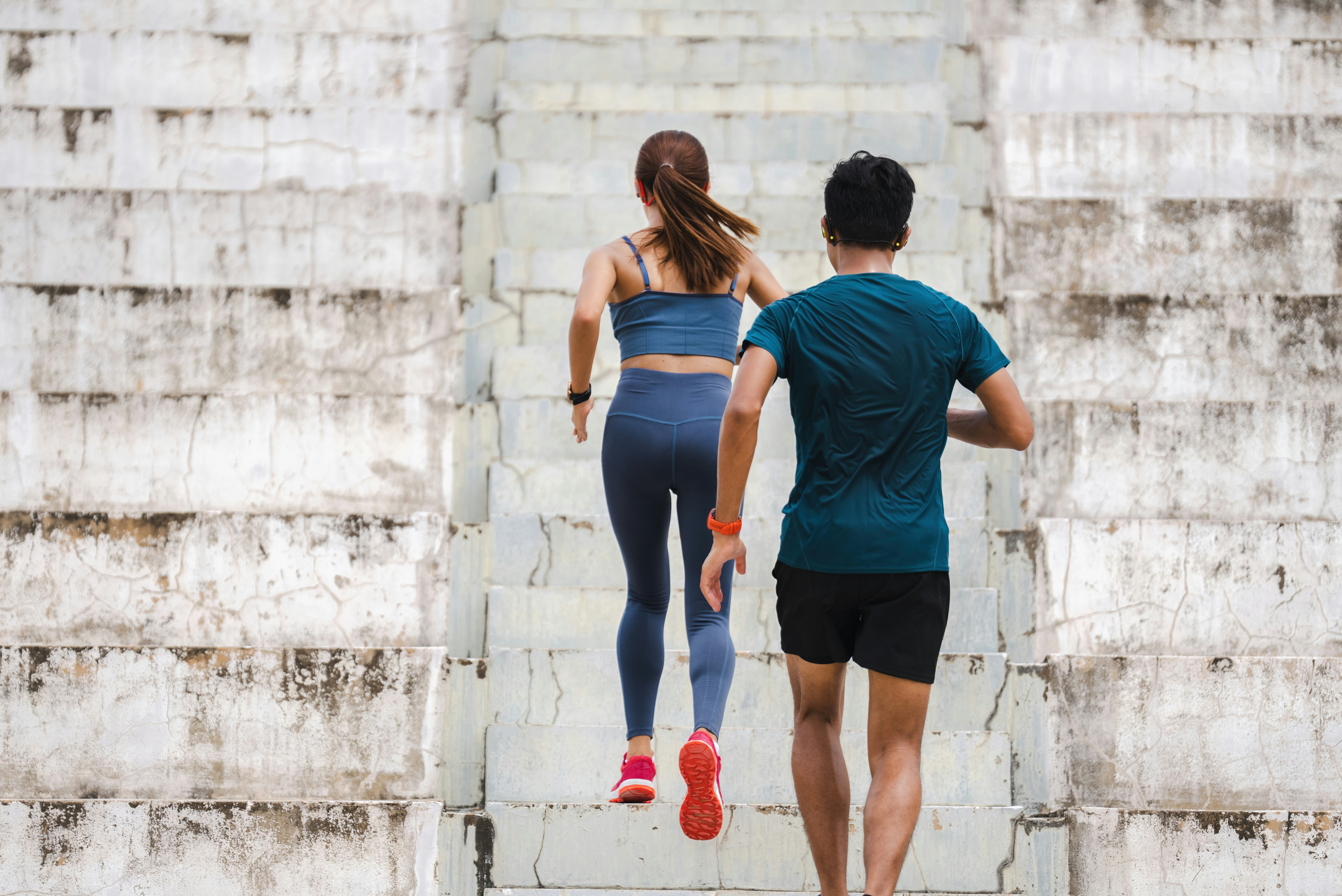 Two people viewed from behind running up a wide weathered concrete stadium staircase, the woman in blue athletic wear and the man in a dark teal shirt, on an overcast day