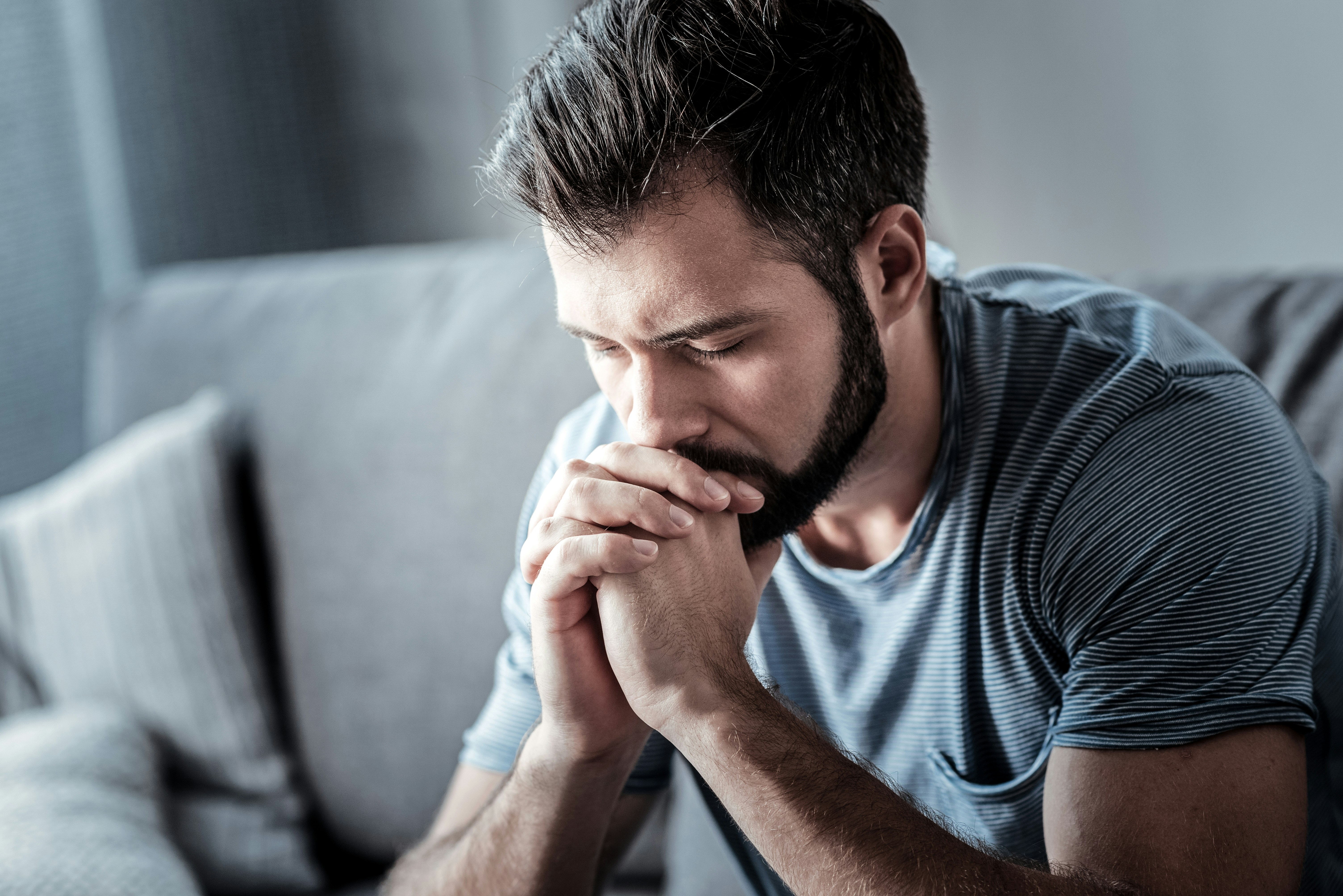 A young man with a dark beard sitting on a grey sofa with eyes closed and hands clasped in front of his face, wearing a blue-and-white striped t-shirt, in a soft contemplative mood