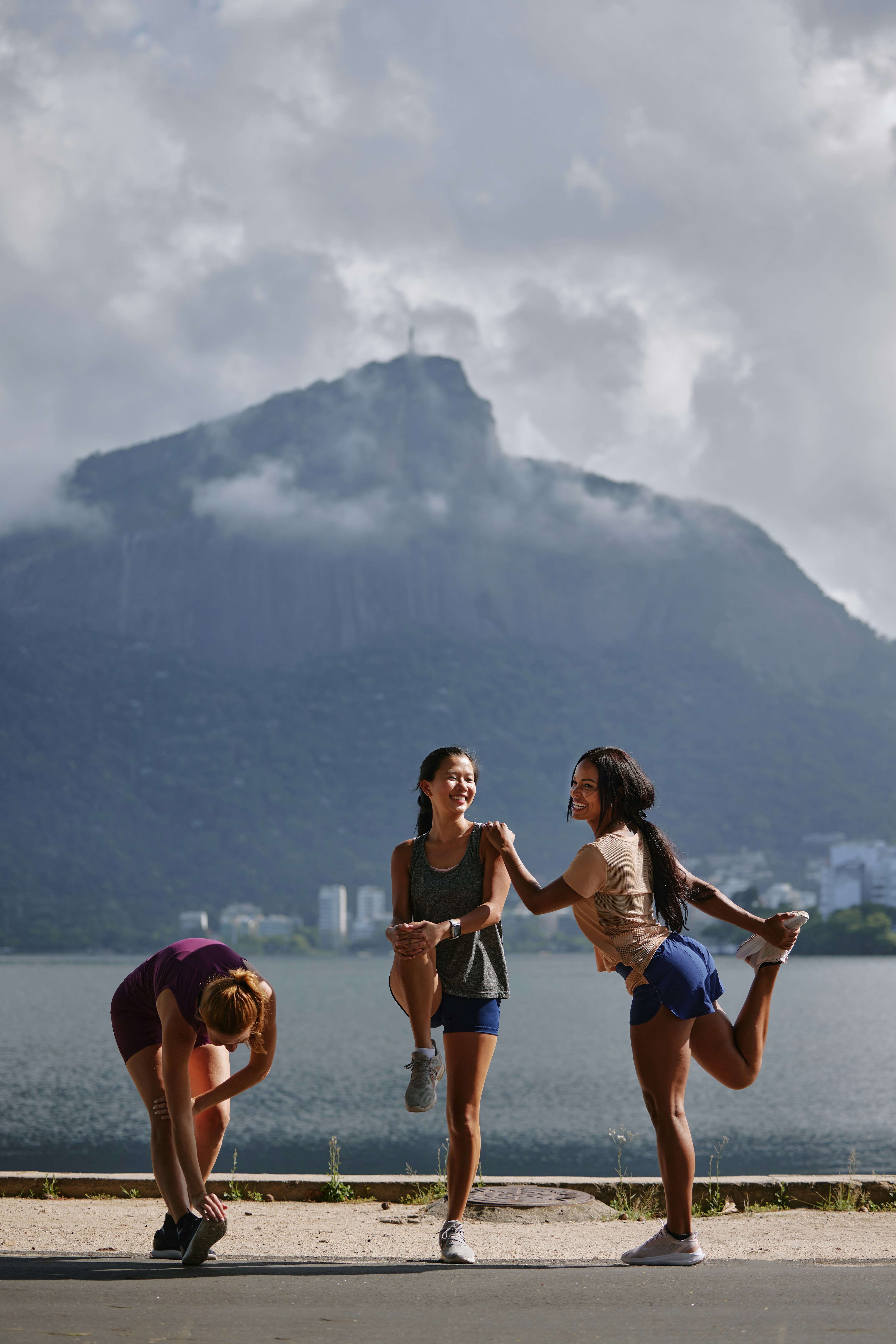 Three athletic women performing warm-up stretches outdoors on a waterfront surface, one balancing on one leg and two doing quad and hamstring stretches, with a large mountain in the hazy background