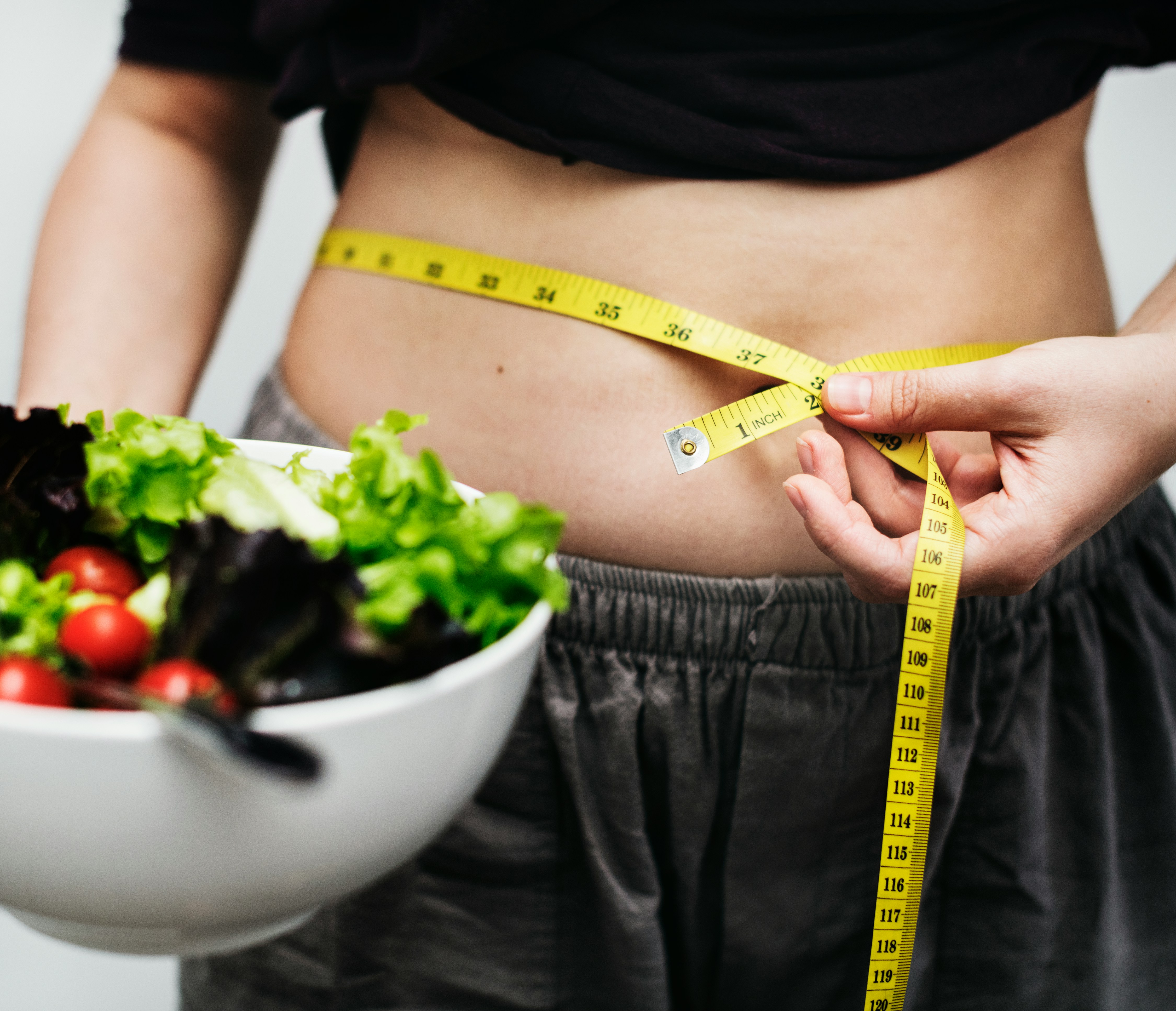 A person in a dark top and grey shorts holding a white bowl of green salad with cherry tomatoes, with a yellow measuring tape wrapped around their bare abdomen