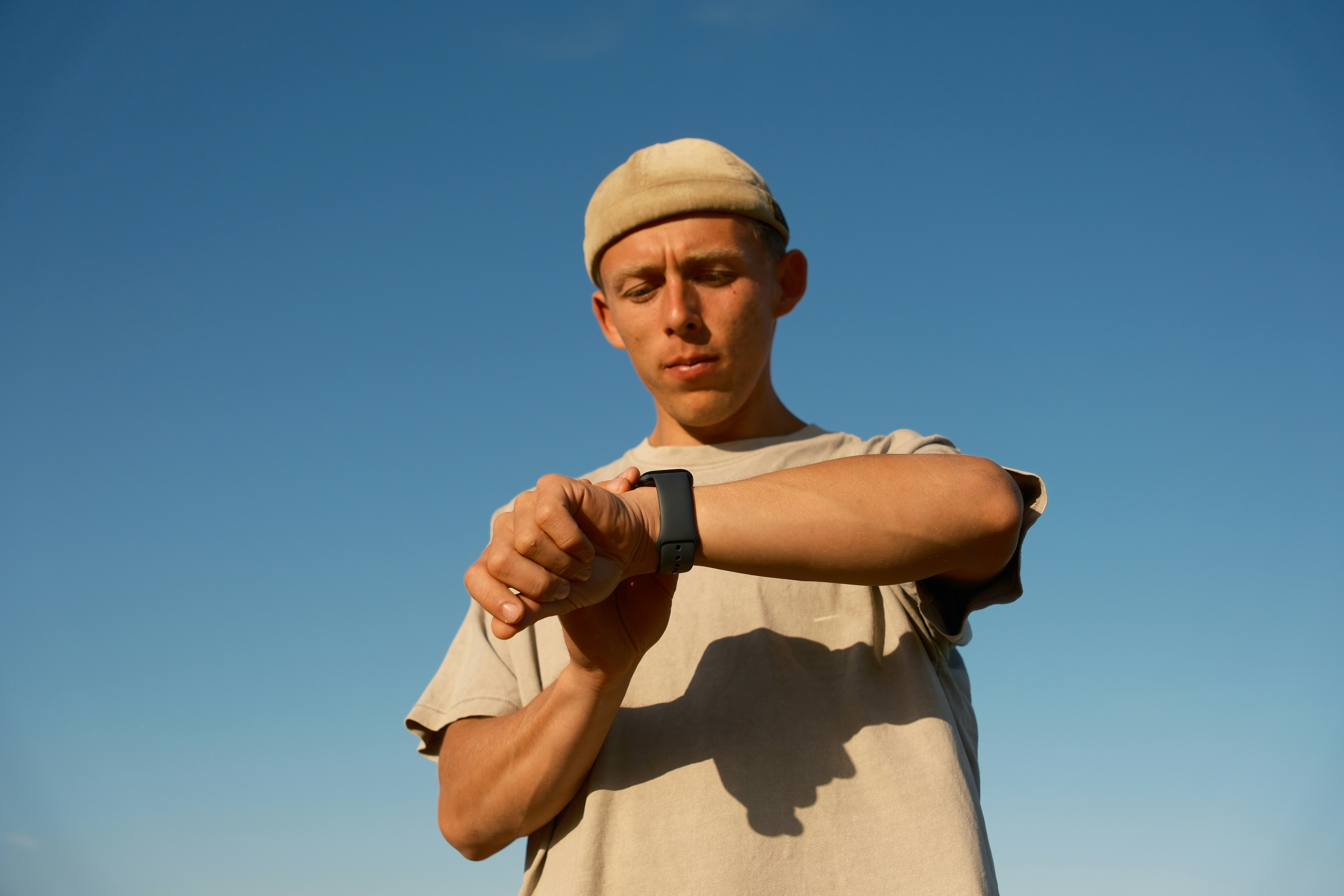 A young man in a beige t-shirt and beanie looking down at his left wrist to check a smartwatch, against a bright blue sky