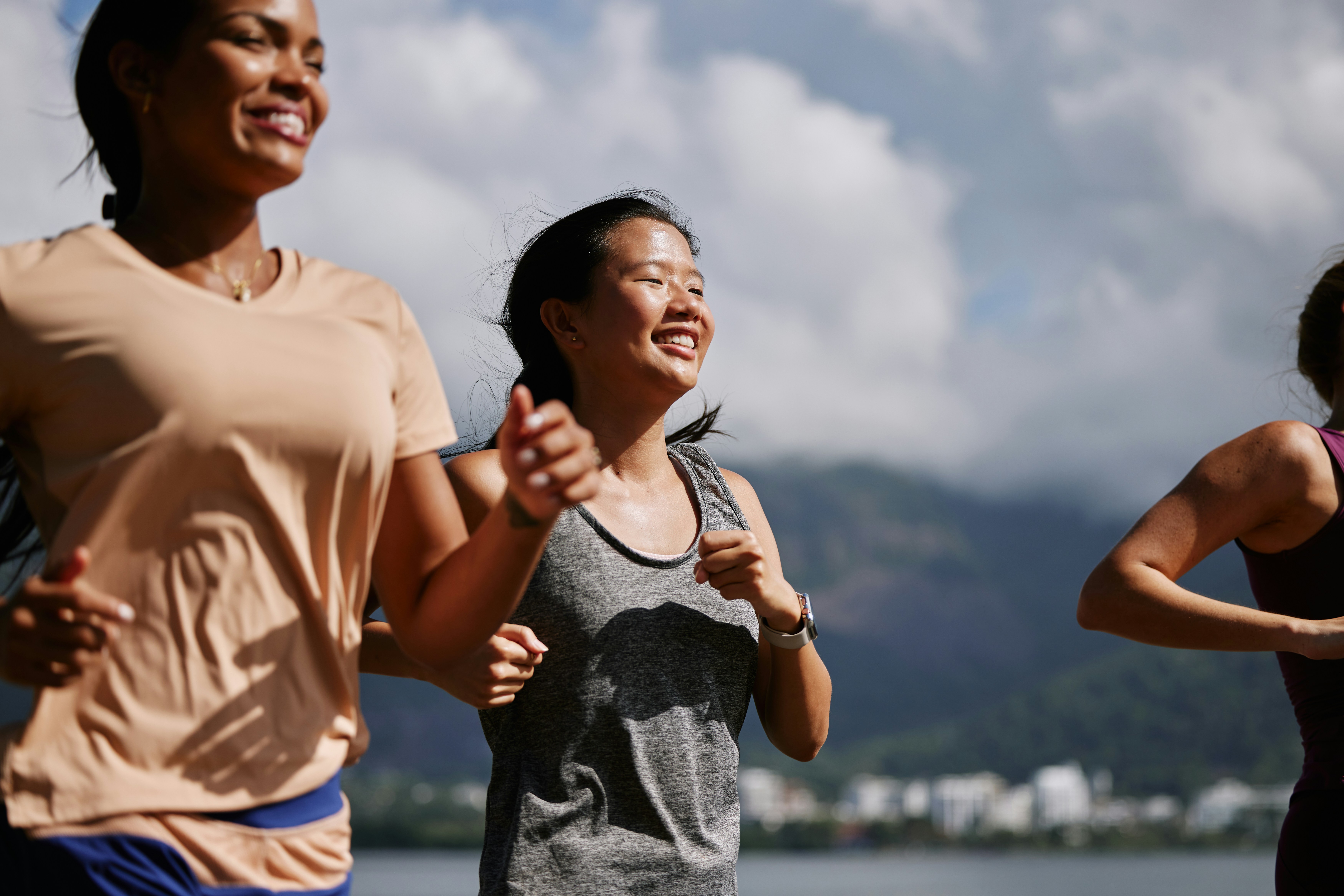 Three women jogging together outdoors at a side profile, the foreground runner in a grey tank top smiling broadly, against a blurred mountain and waterfront background