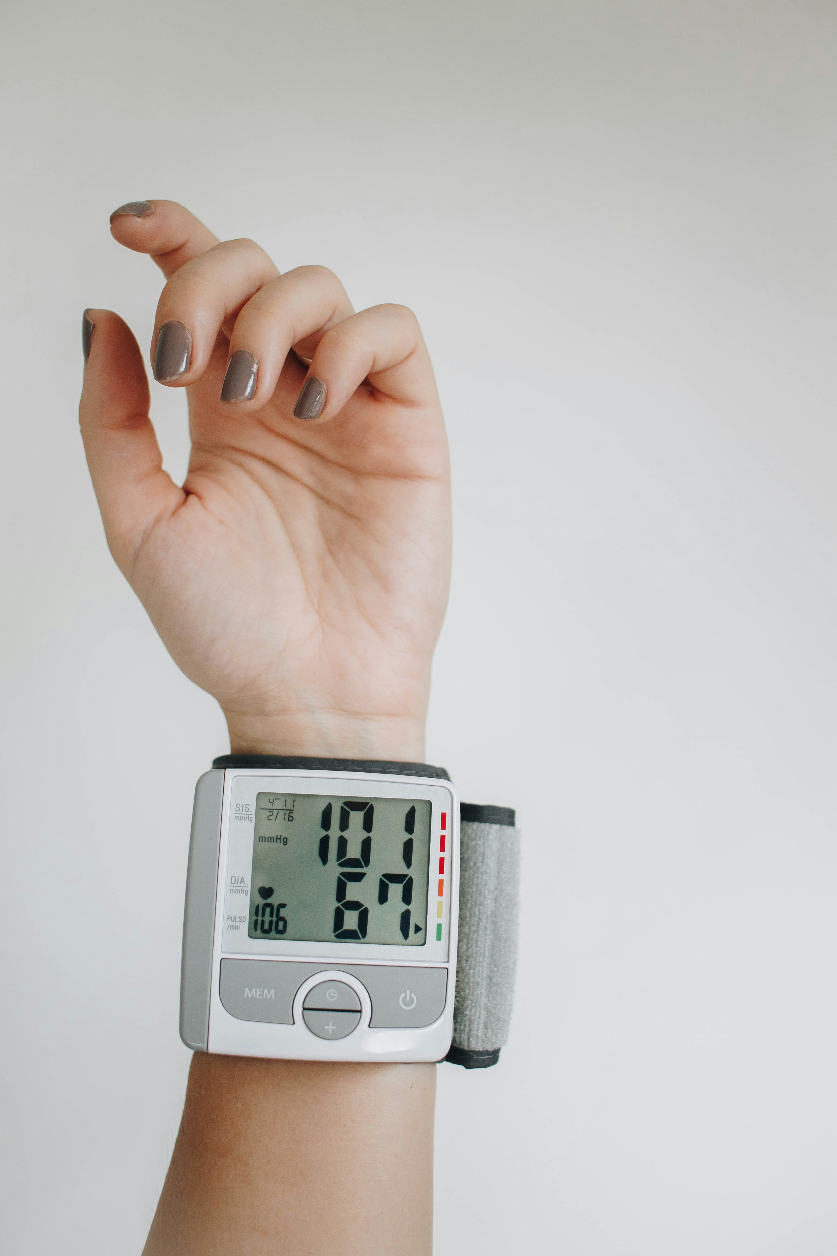 A close-up of a hand wearing a digital wrist blood pressure monitor displaying systolic, diastolic, and pulse readings with a color-coded indicator bar, against an off-white background