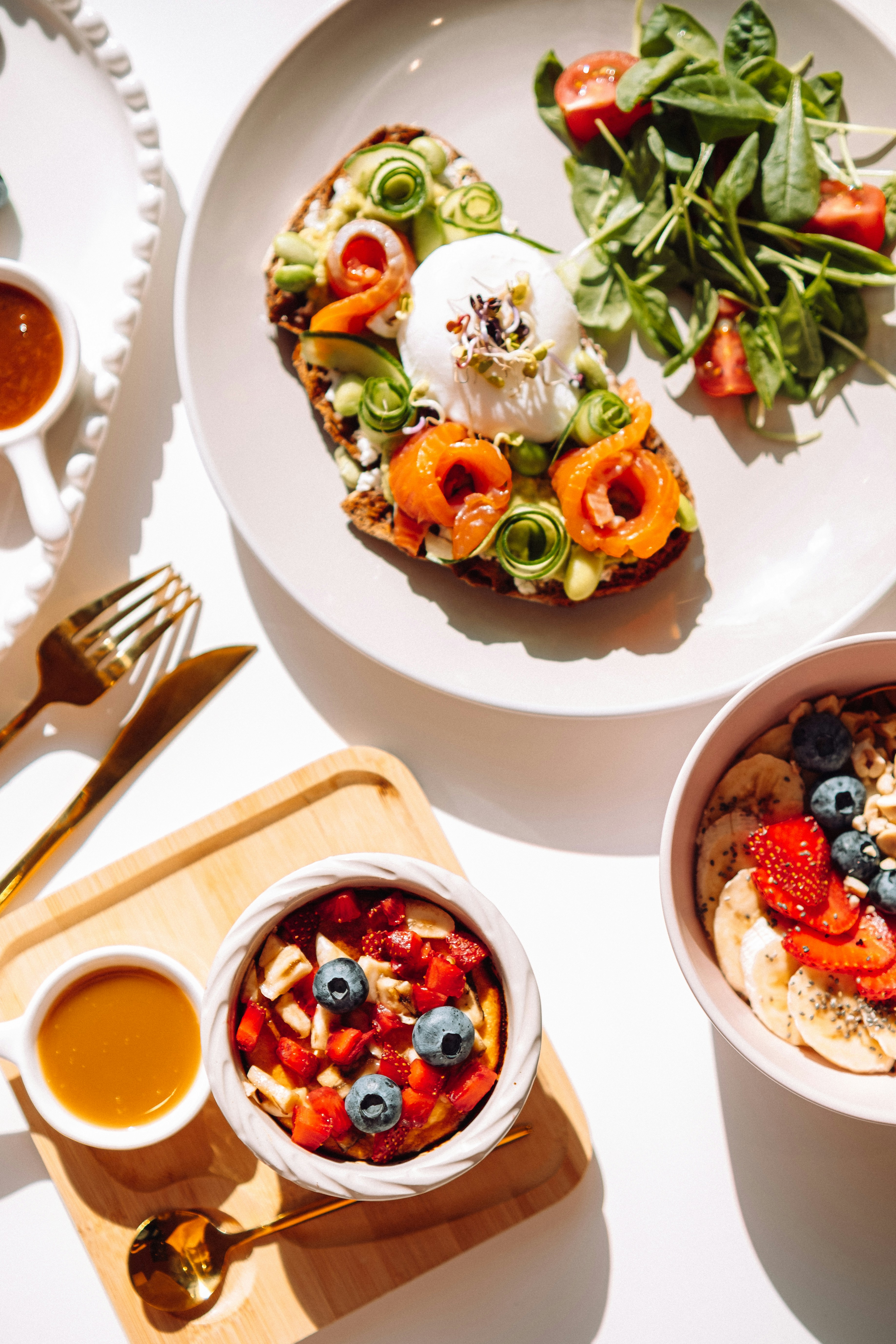 An overhead top-down view of a healthy breakfast spread on a white surface with avocado toast and smoked salmon, a smoothie bowl, and porridge with fresh fruit, in sharp natural light