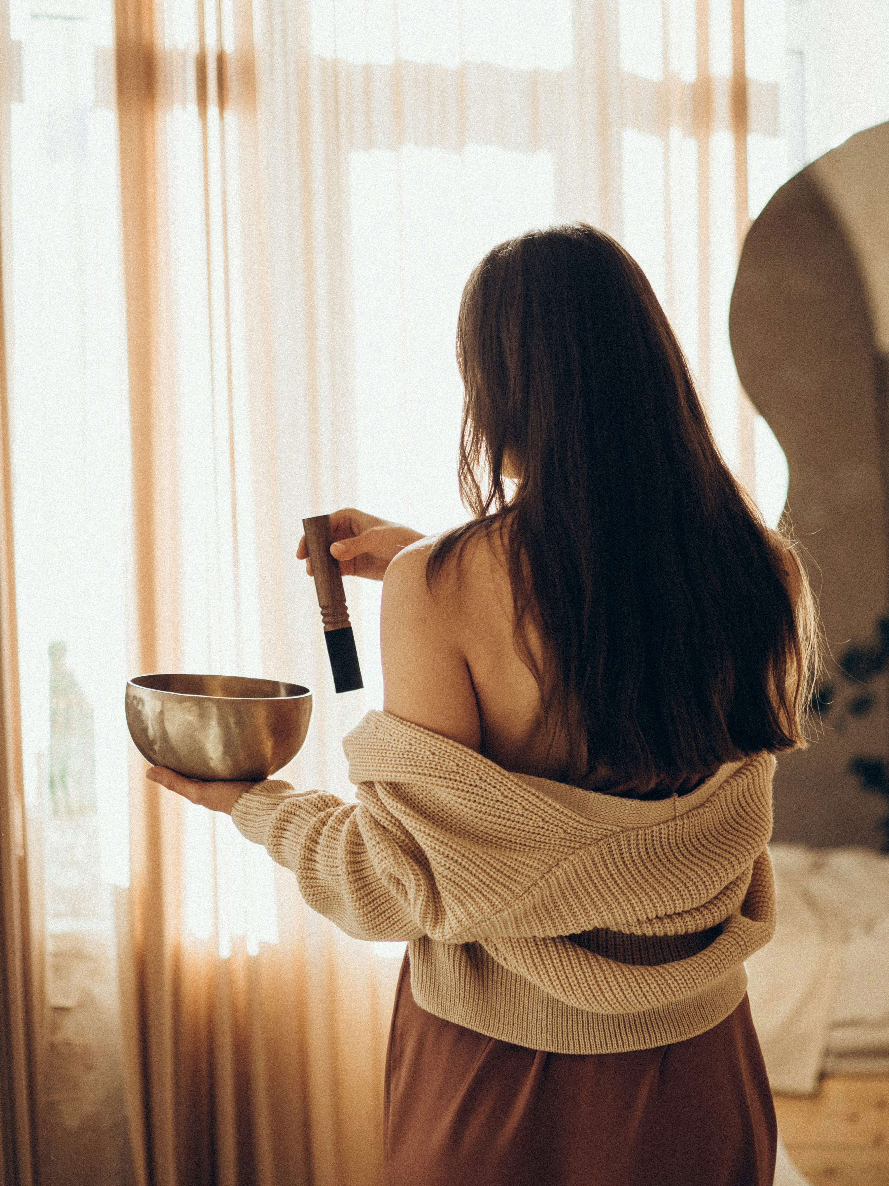A woman with long dark hair viewed from behind, holding a gold singing bowl and wooden mallet in front of a window with sheer beige curtains, in a calm meditative setting