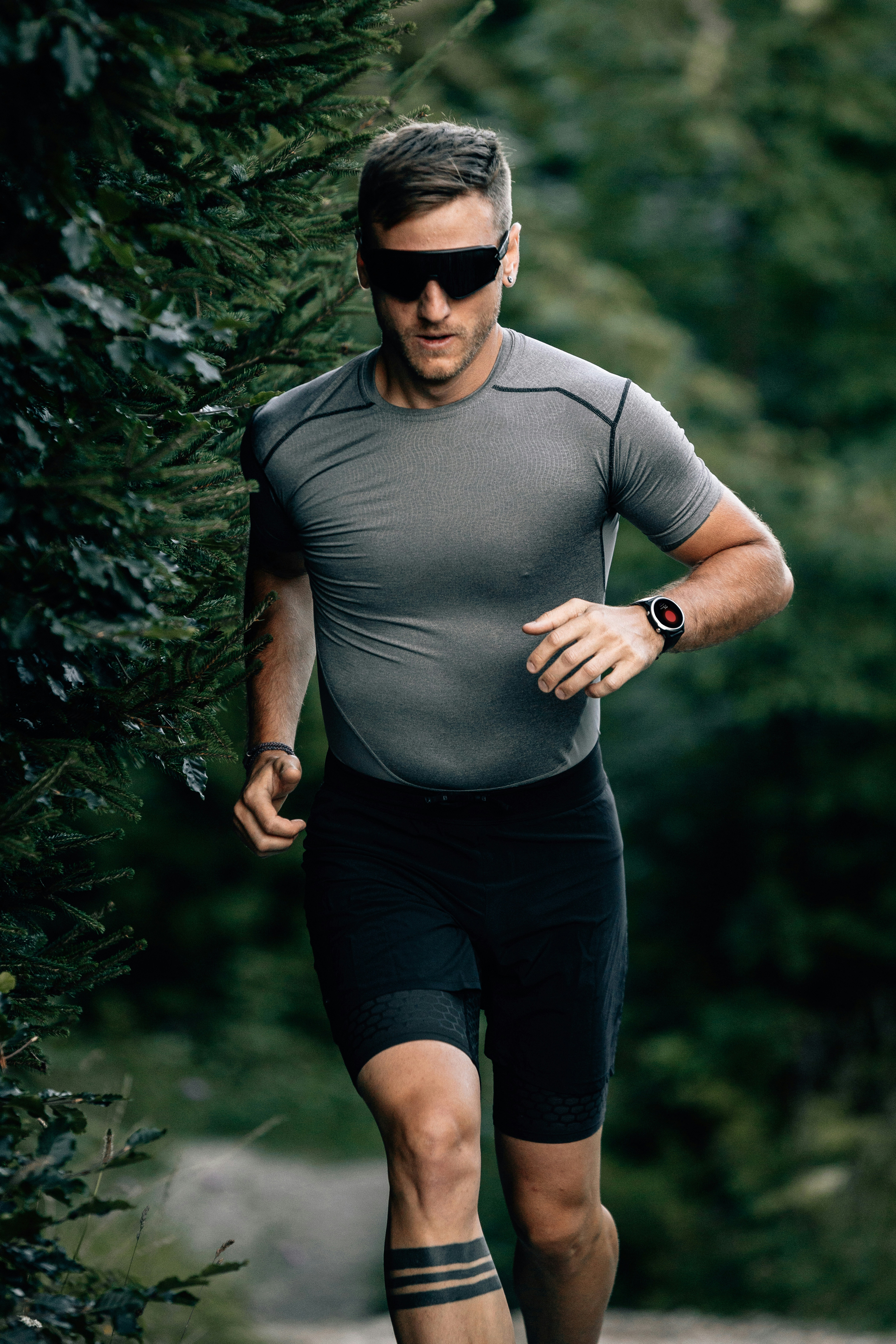 A male runner in a charcoal athletic shirt and black shorts running toward the camera on a forest trail, wearing dark wrap-around sunglasses and a fitness watch, with soft-focus coniferous trees behind