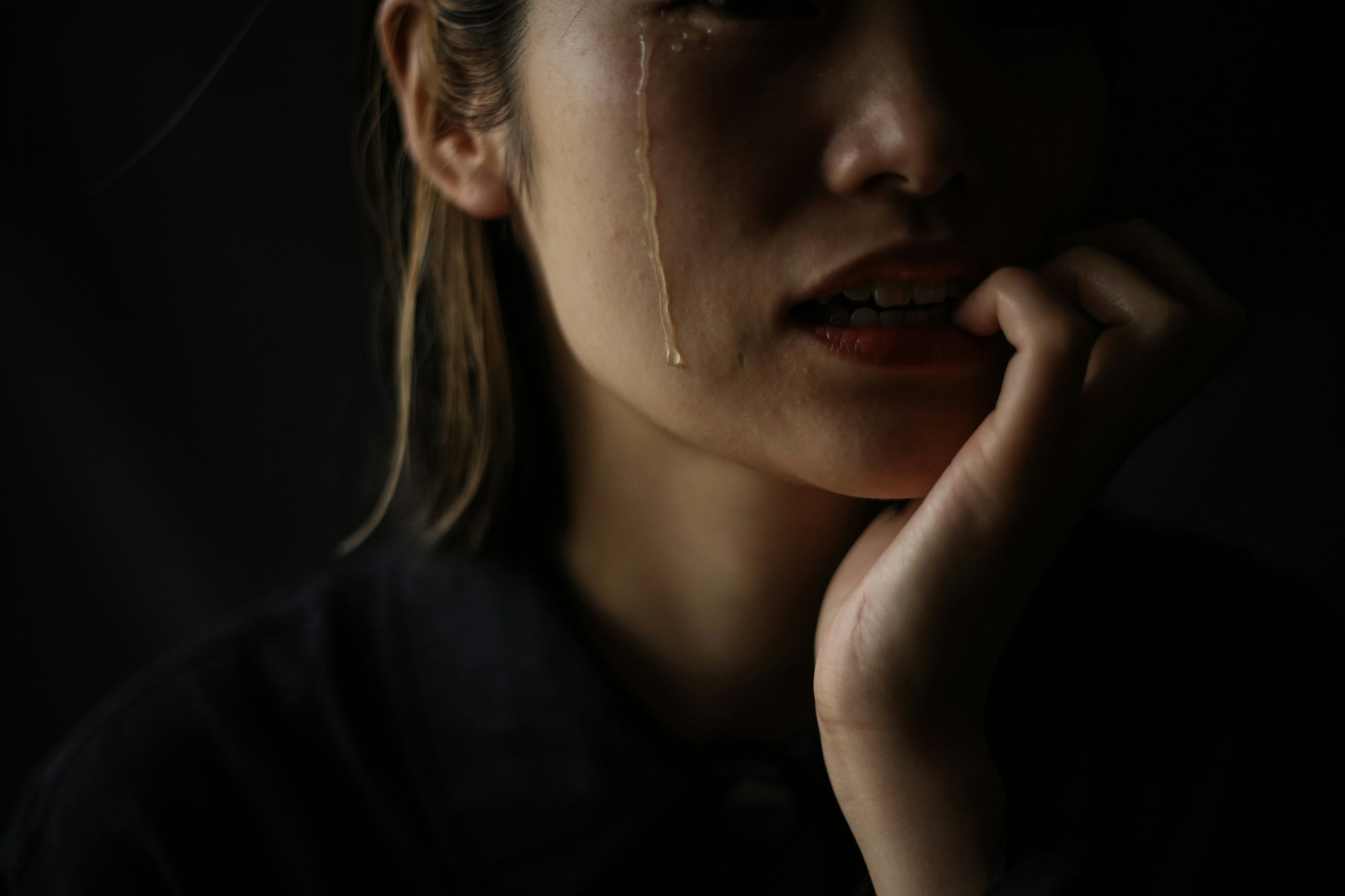 A close-up of a person's lower face with a single tear tracking down the cheek, lips parted and biting the lower lip, one hand raised to the mouth, in dramatic low-key lighting against a dark background