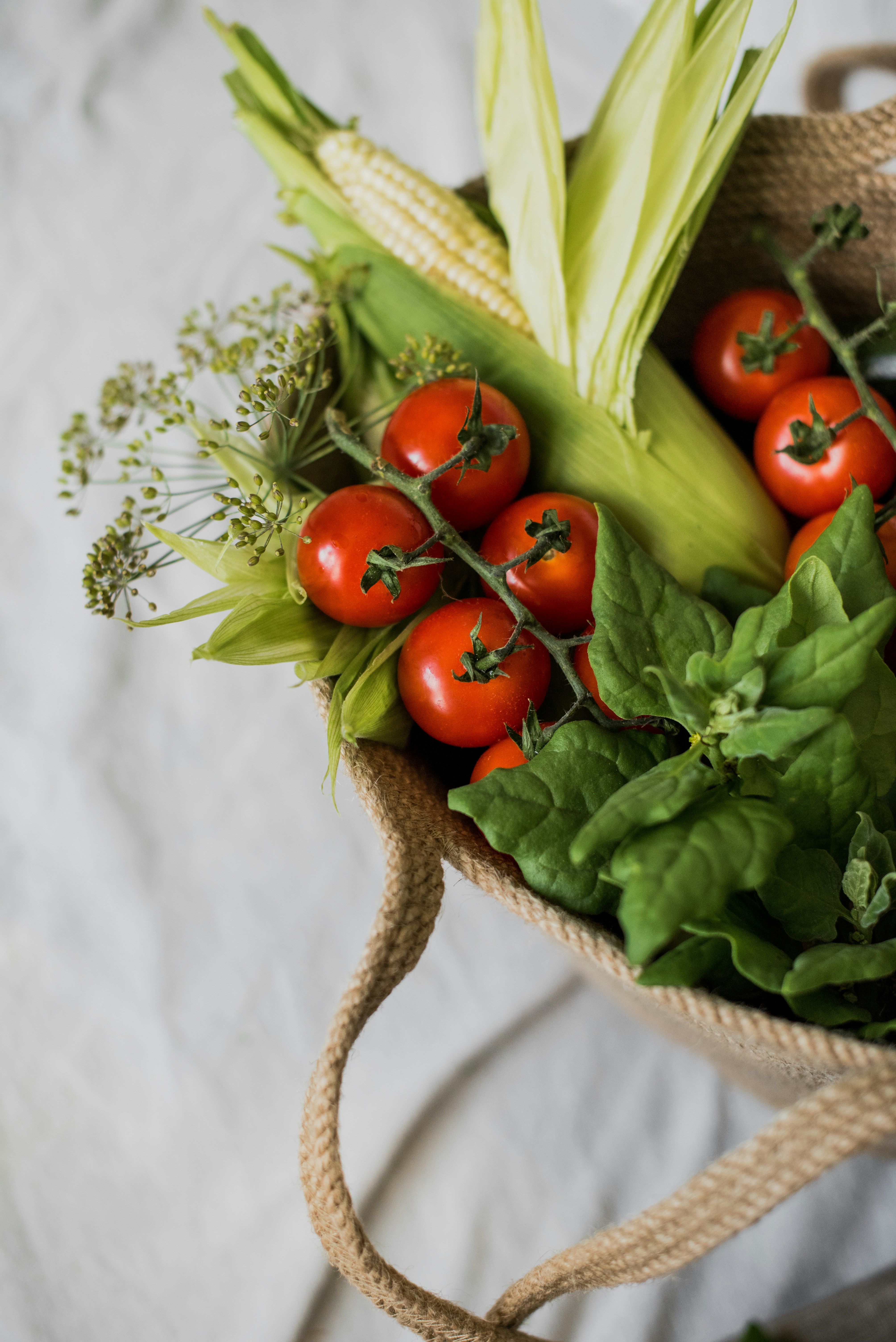 A top-down view of a woven jute market bag filled with cherry tomatoes on the vine, corn in husks, dark leafy greens, and a sprig of dill, resting on a soft white fabric surface