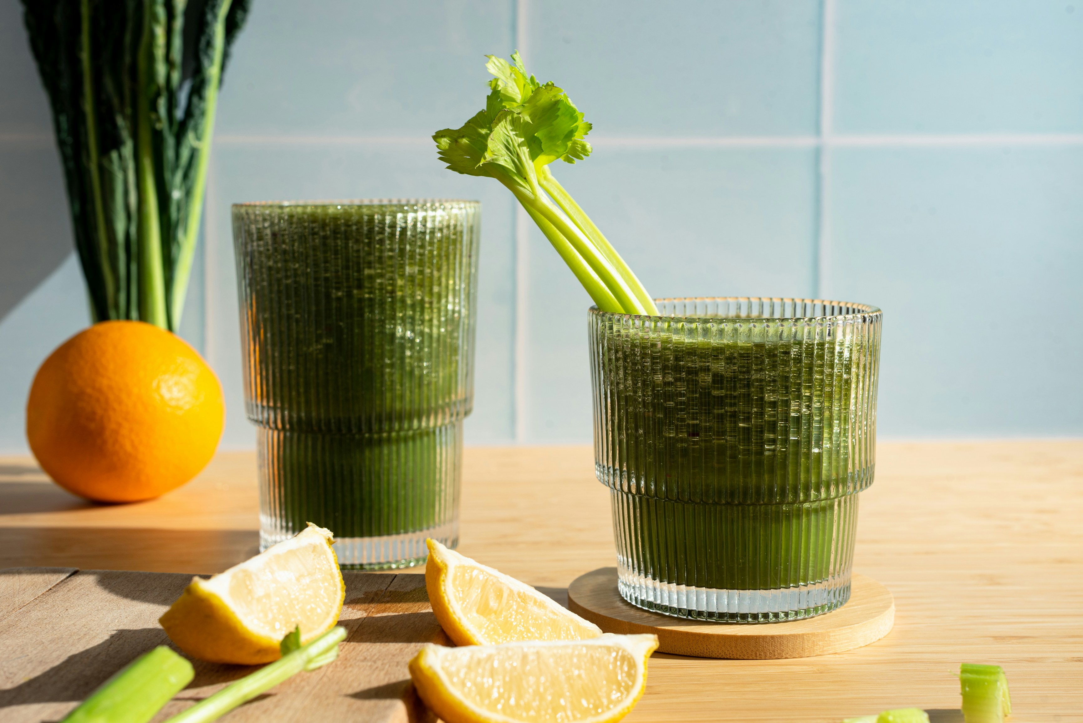 Two ribbed glass tumblers filled with dark green smoothie on a light wooden surface, one with a celery stalk protruding, flanked by an orange, kale, and lemon wedges against a pale blue tiled wall