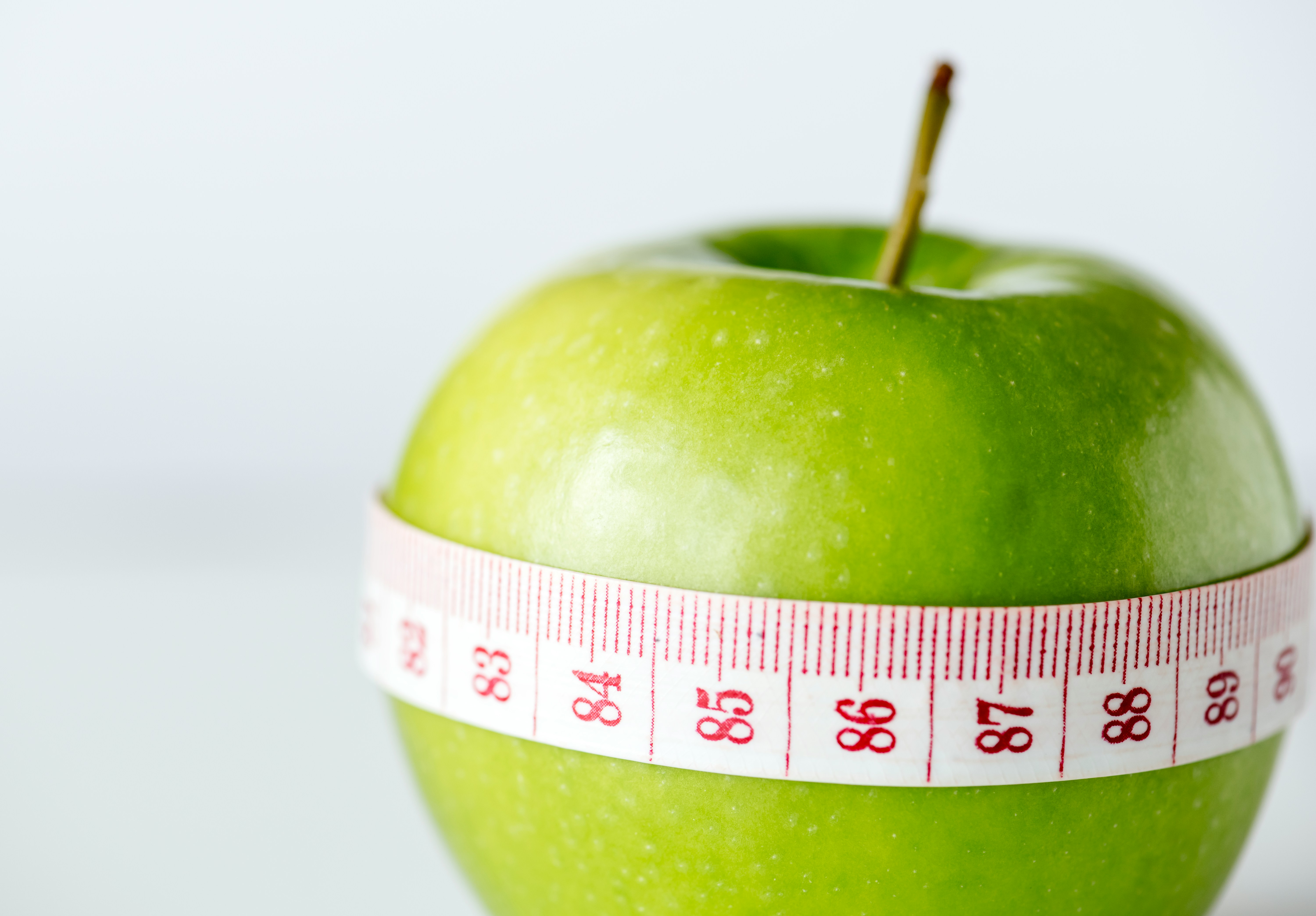 A single bright green apple wrapped horizontally with a white measuring tape showing measurement markings, against a soft off-white background
