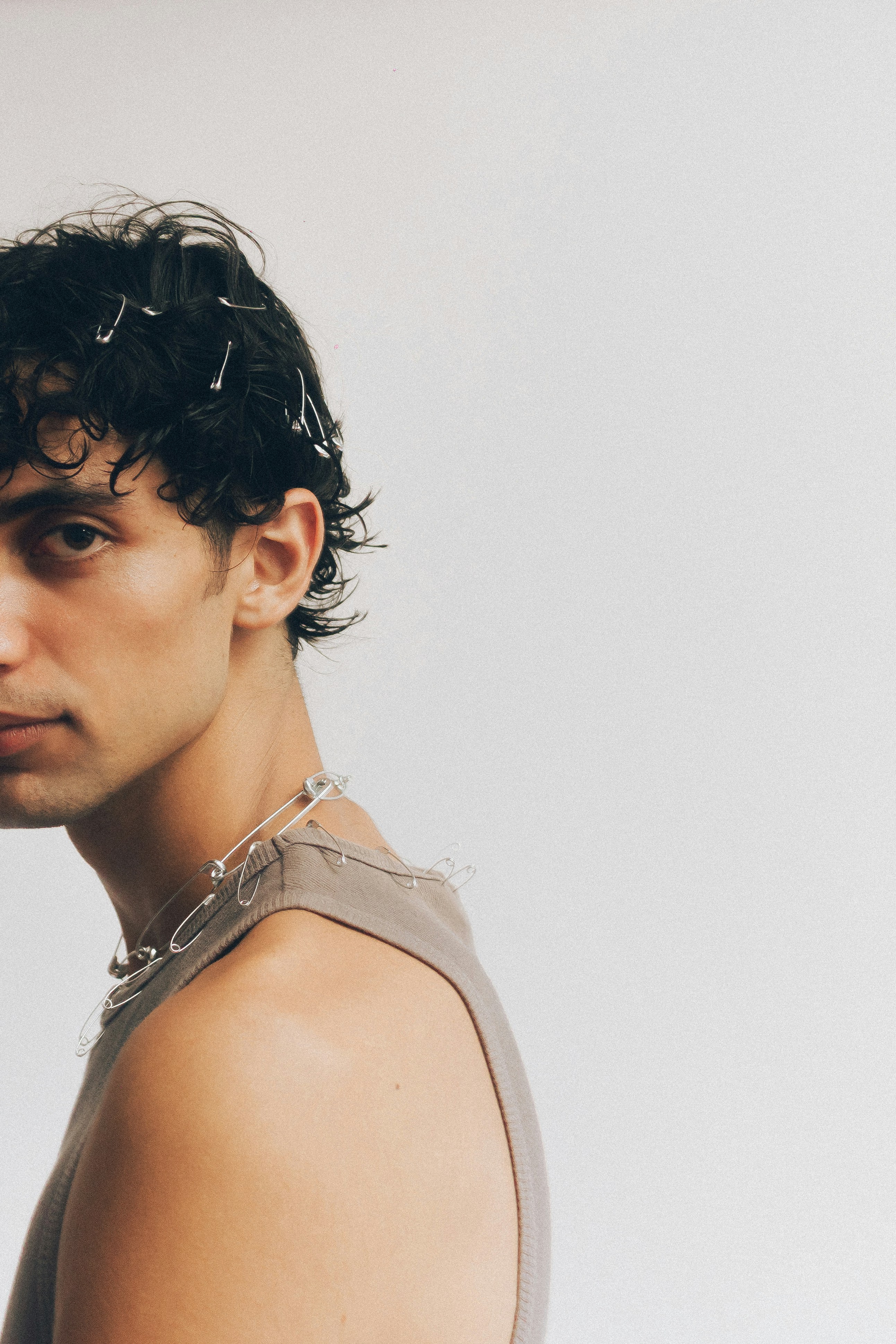 A young man with dark wavy hair styled with metallic safety pins, wearing a taupe ribbed tank top and a safety-pin necklace, against a light grey background
