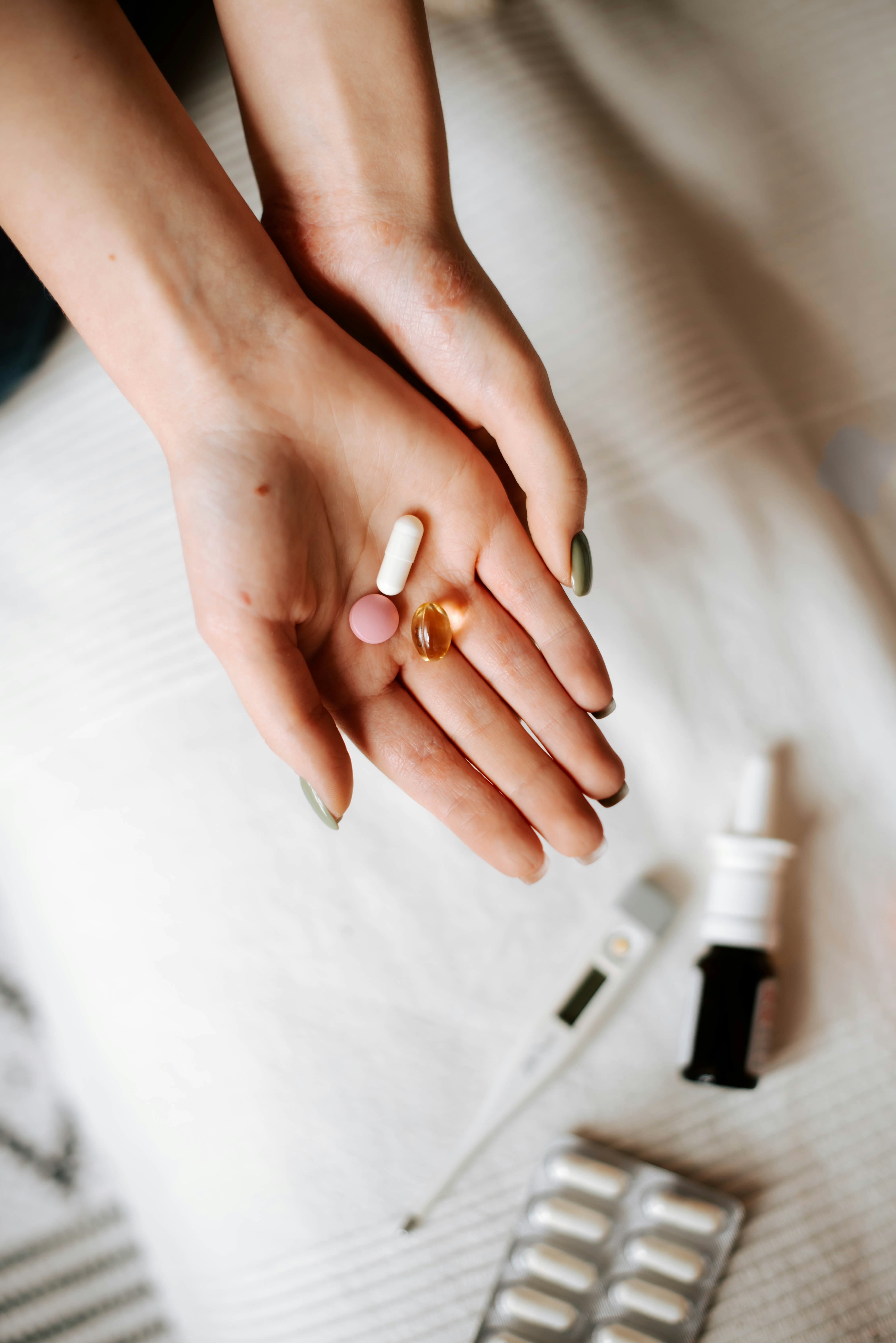 A top-down view of cupped hands holding a white capsule, a pink tablet, and a golden softgel, with a digital thermometer, a dark glass nasal spray bottle, and a foil blister pack on a cream fabric surface