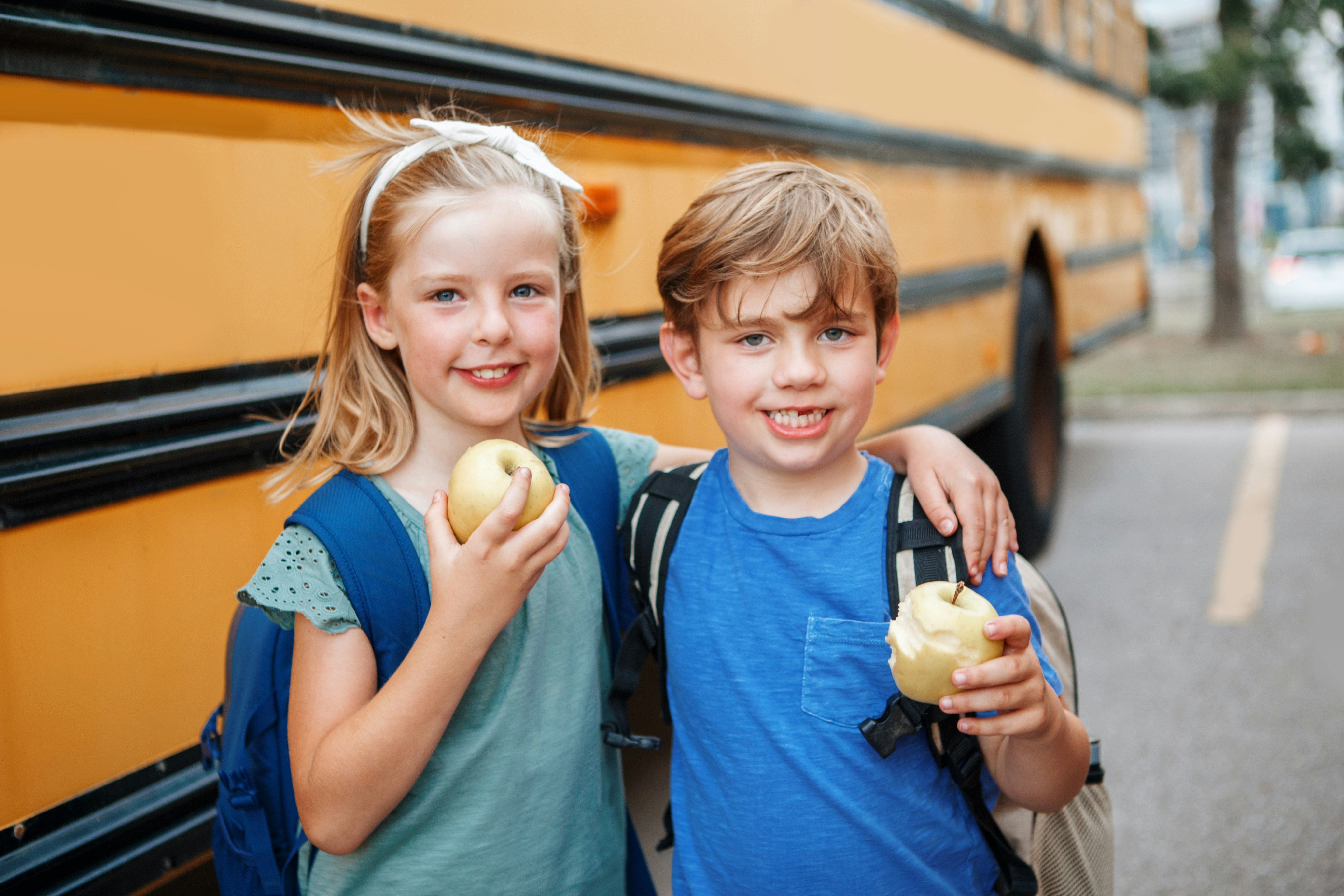 Two children standing side by side in front of a yellow school bus, each holding a yellow apple, the girl in a mint-green top and the boy in a blue t-shirt smiling