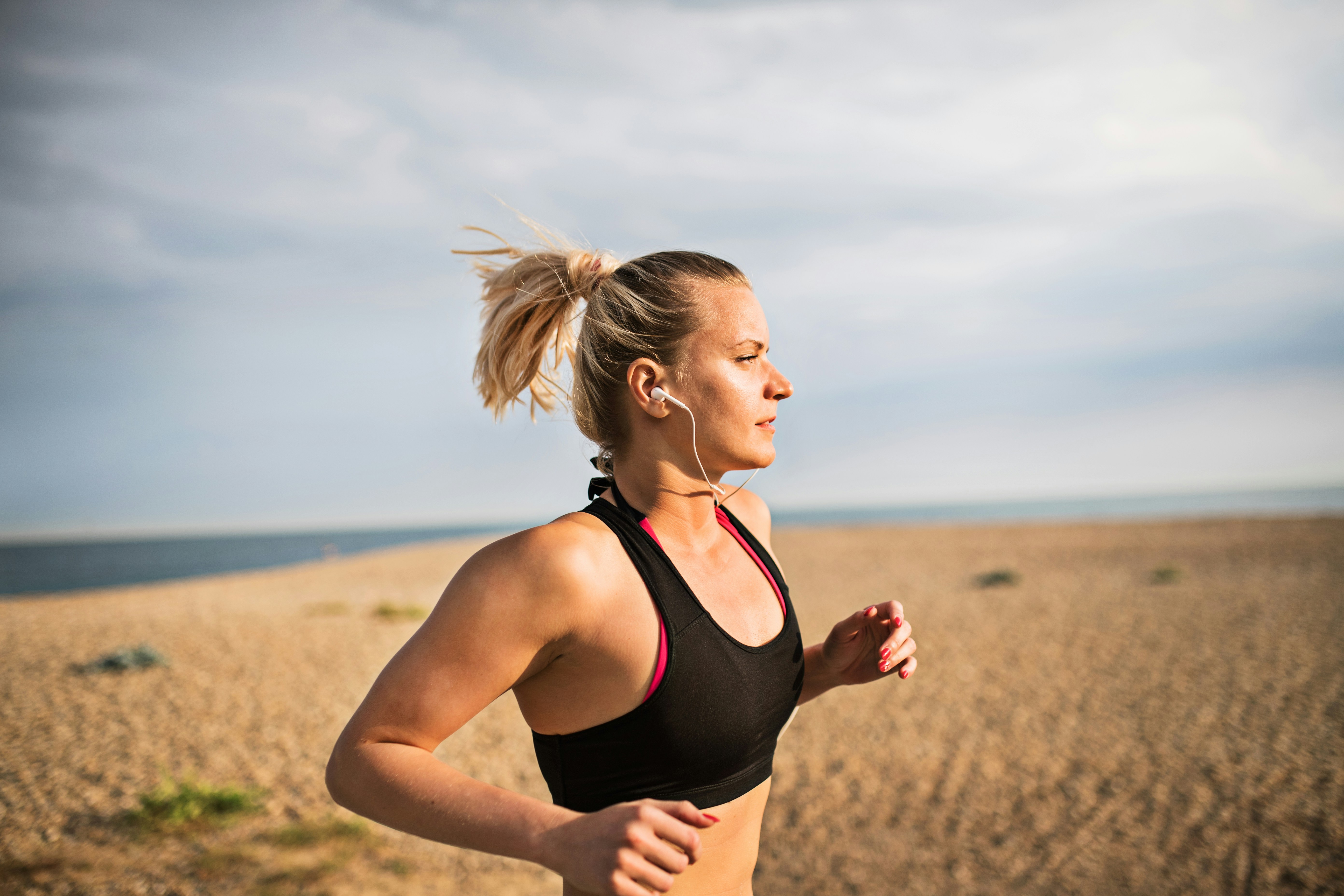 A woman with blonde hair in a ponytail running in profile on a sandy beach beside the sea, wearing a black sports bra, in soft natural light