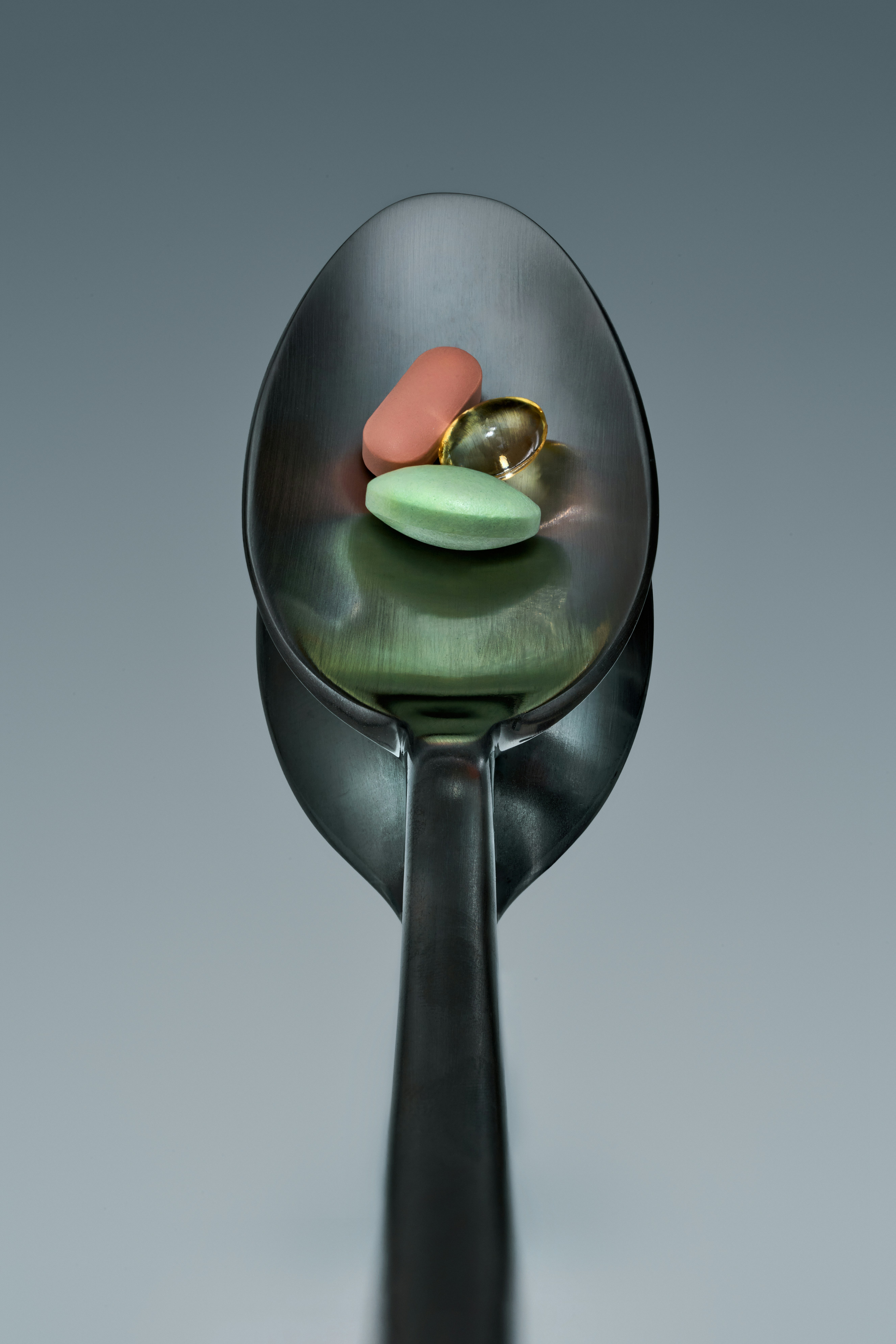 Three supplement pills — a peach tablet, a golden softgel, and a light-green tablet — resting in a stainless steel spoon against a smooth neutral gray background