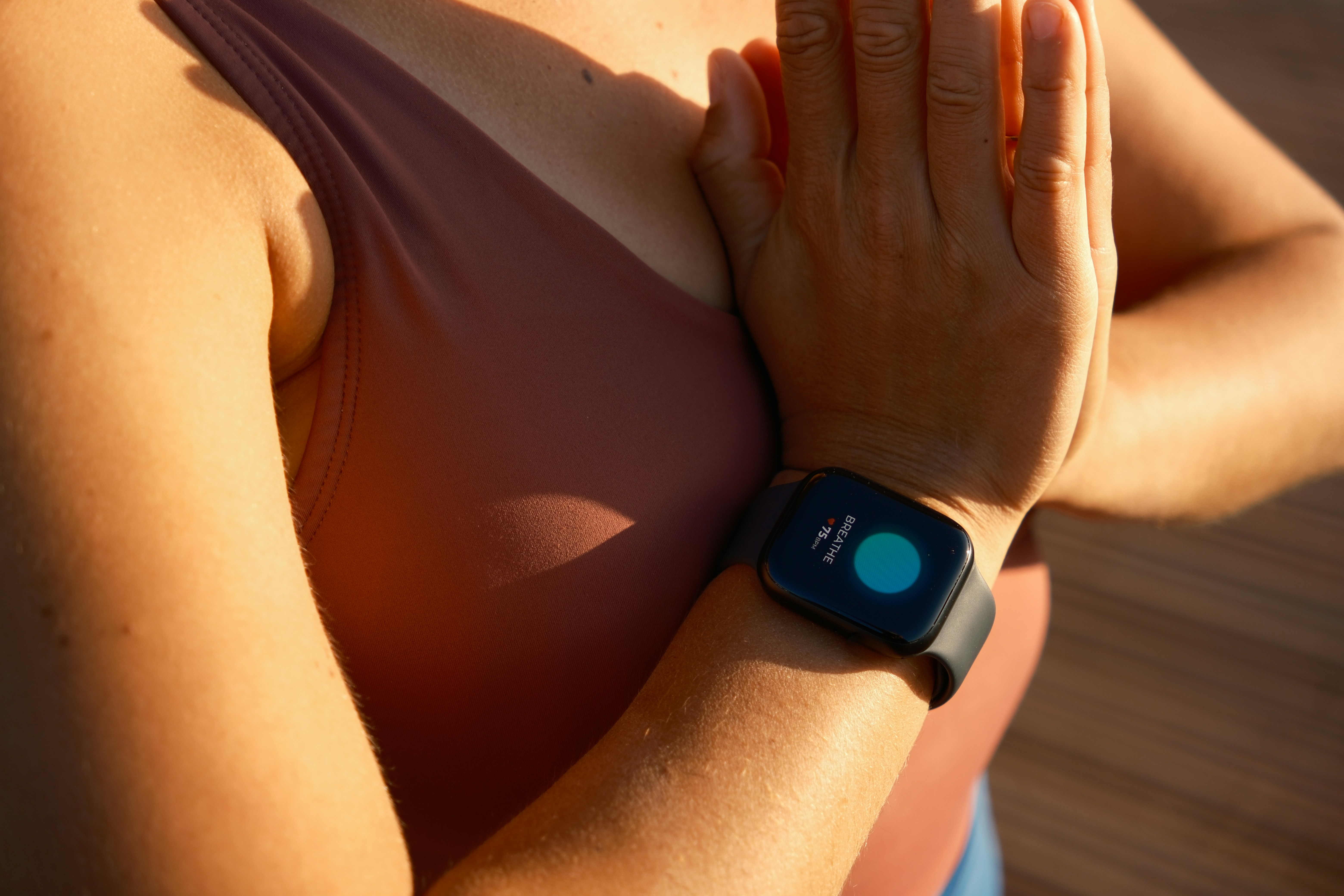 A person's hands pressed together in a prayer position wearing a smartwatch displaying a teal BREATHE interface with 75 BPM, in warm golden-hour light