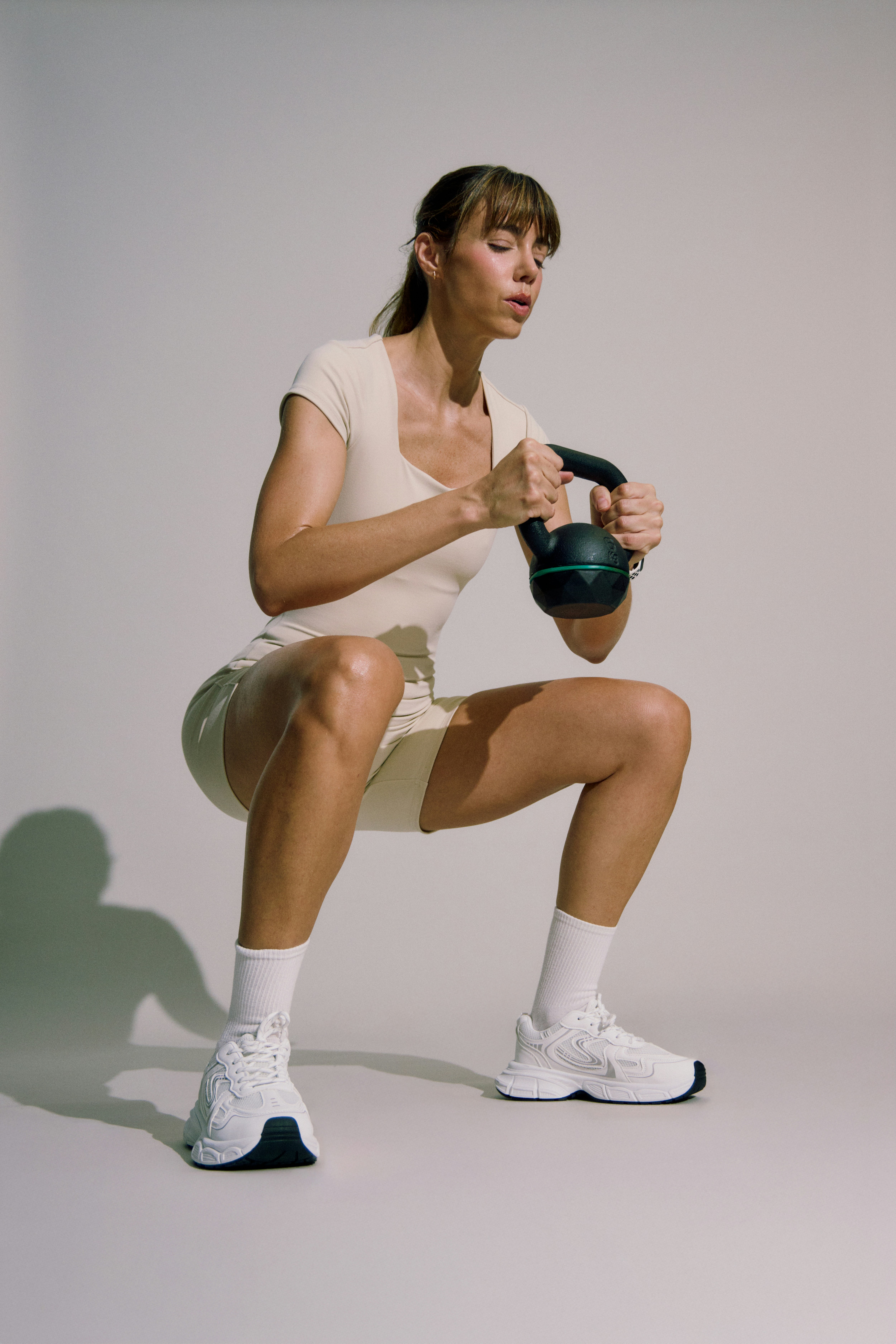 A female athlete in a cream-colored bodysuit performing a deep kettlebell squat holding a black kettlebell at chest height, against a neutral grey studio background with soft lighting