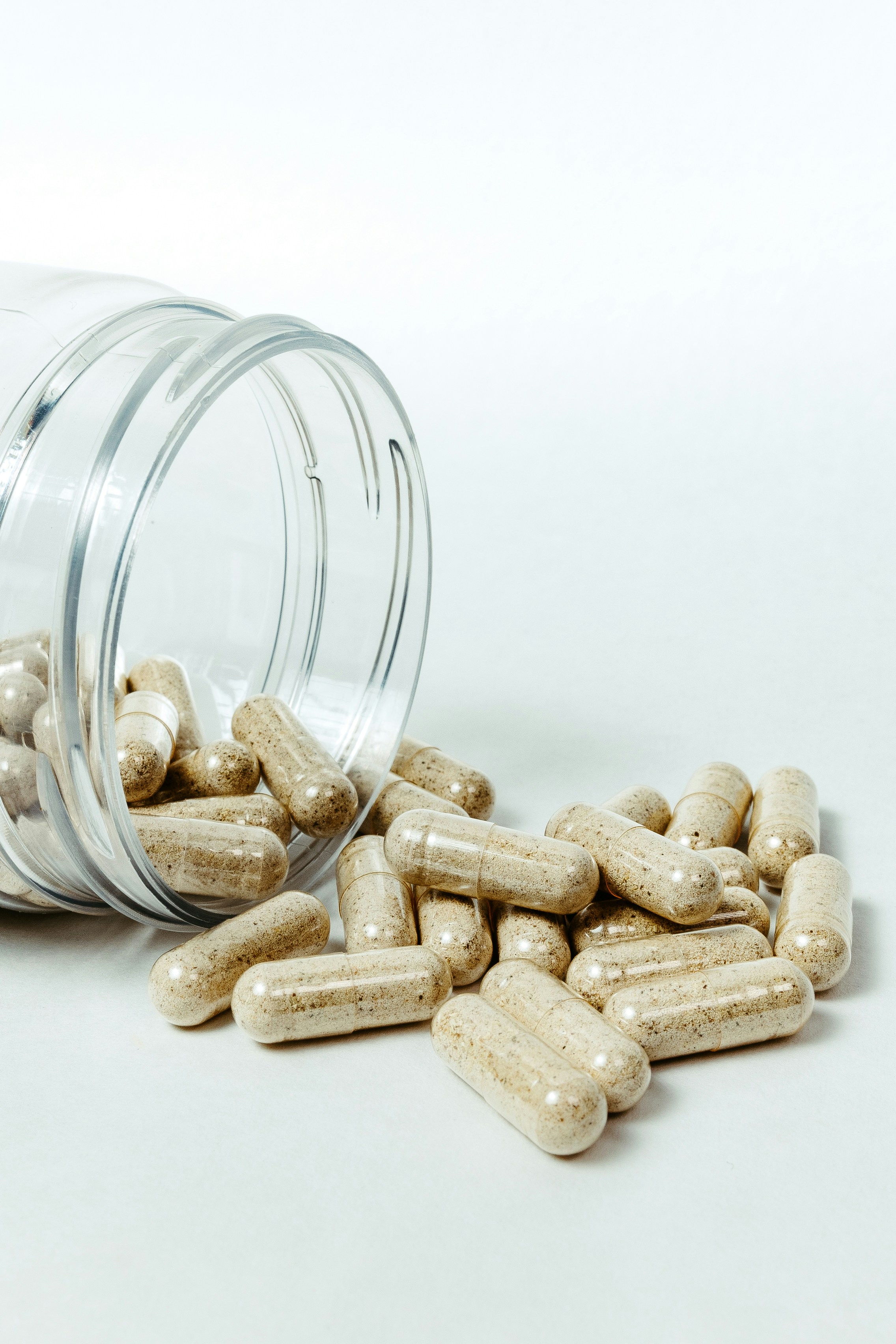 A high-angle close-up of a clear cylindrical plastic jar lying on its side with beige powder-filled capsules spilled out onto an off-white surface in soft directional studio lighting