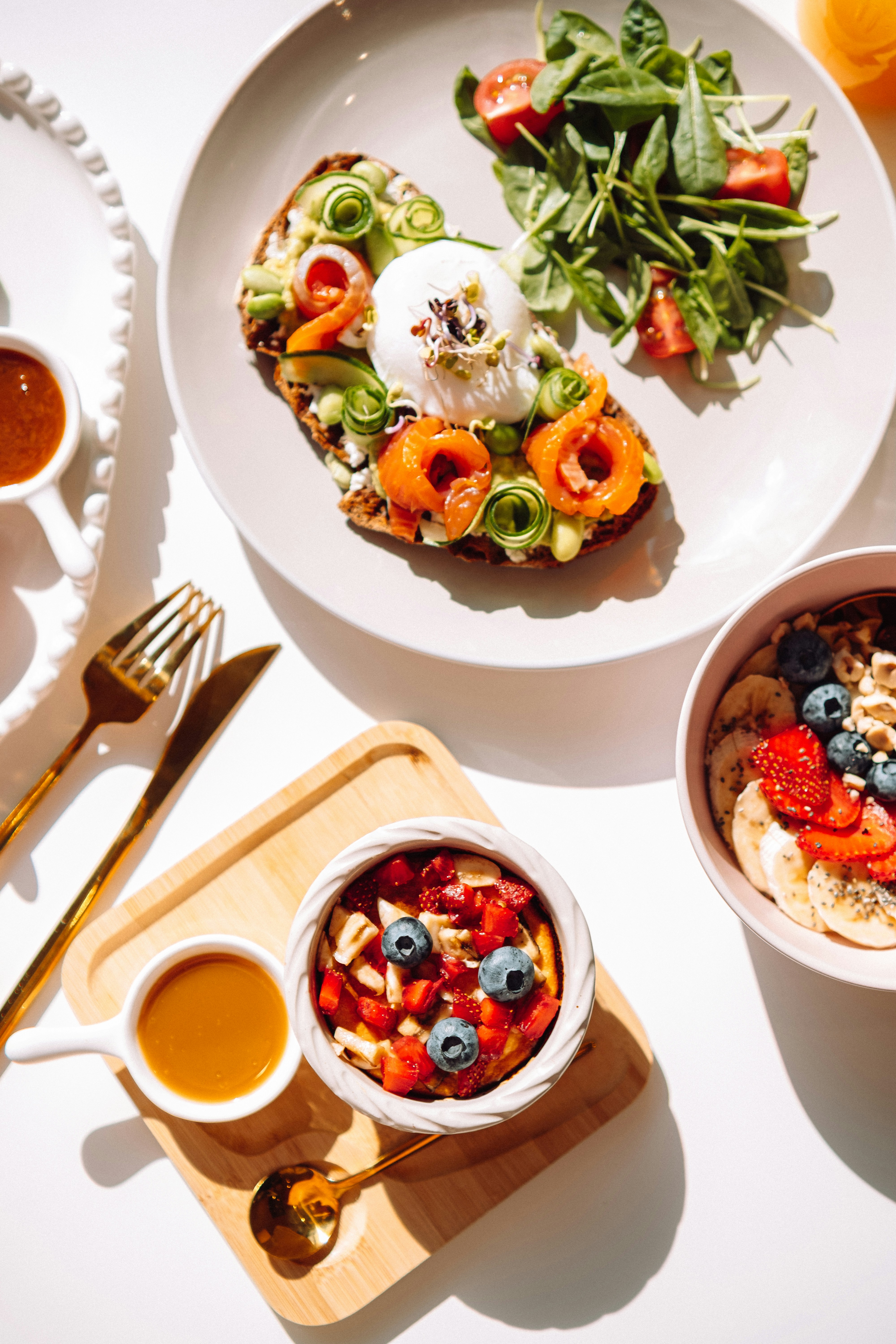 A top-down food flat lay of a healthy meal on a white surface featuring avocado toast with poached egg and smoked salmon, a bowl of fresh berries, and porridge with chia seeds