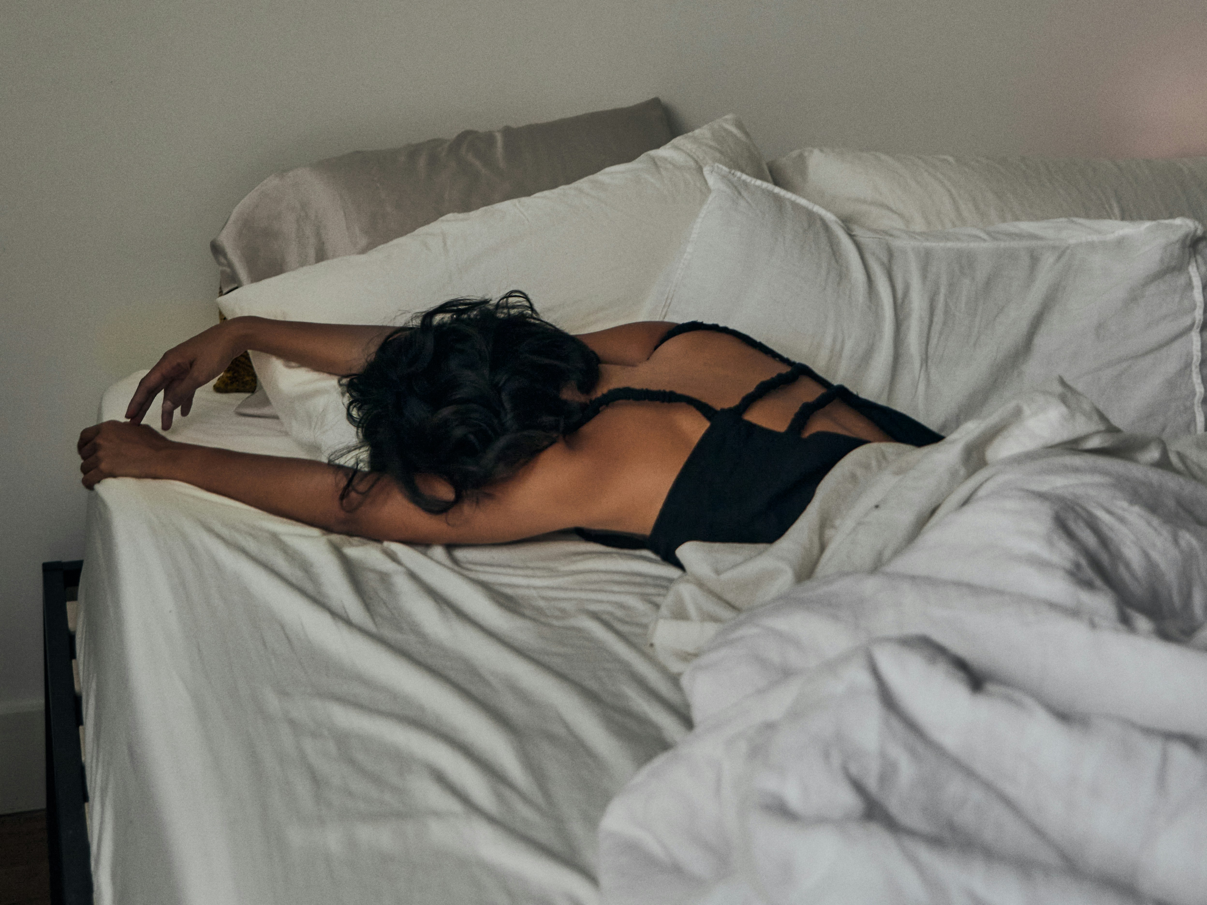 A person with dark wavy hair lying face-down partially under white bed linens, one arm extended, in a calm minimalist bedroom