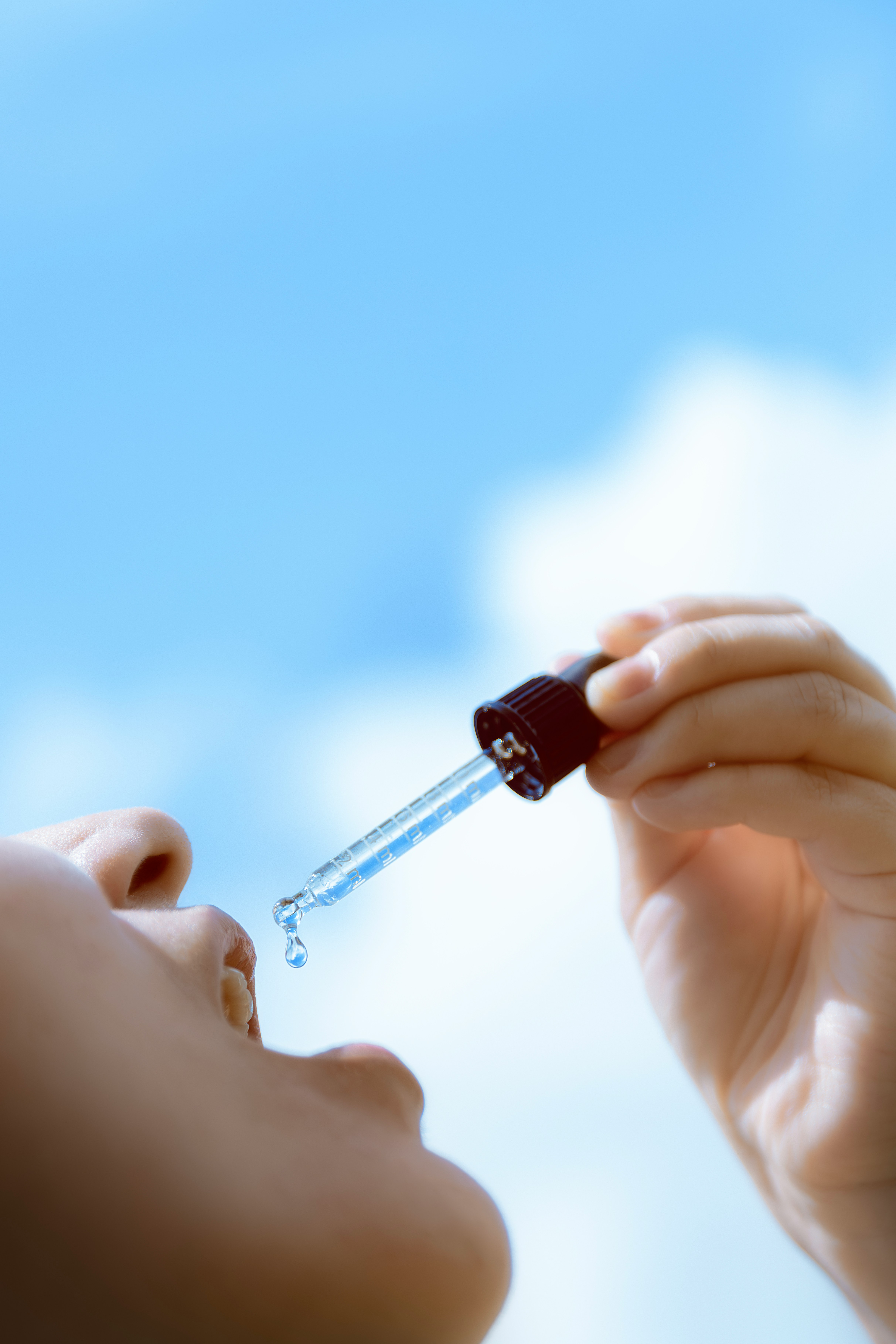 A low-angle close-up of a hand holding a glass medicine dropper with a single drop of clear liquid suspended at the tip above an open mouth, against a bright blurred blue sky