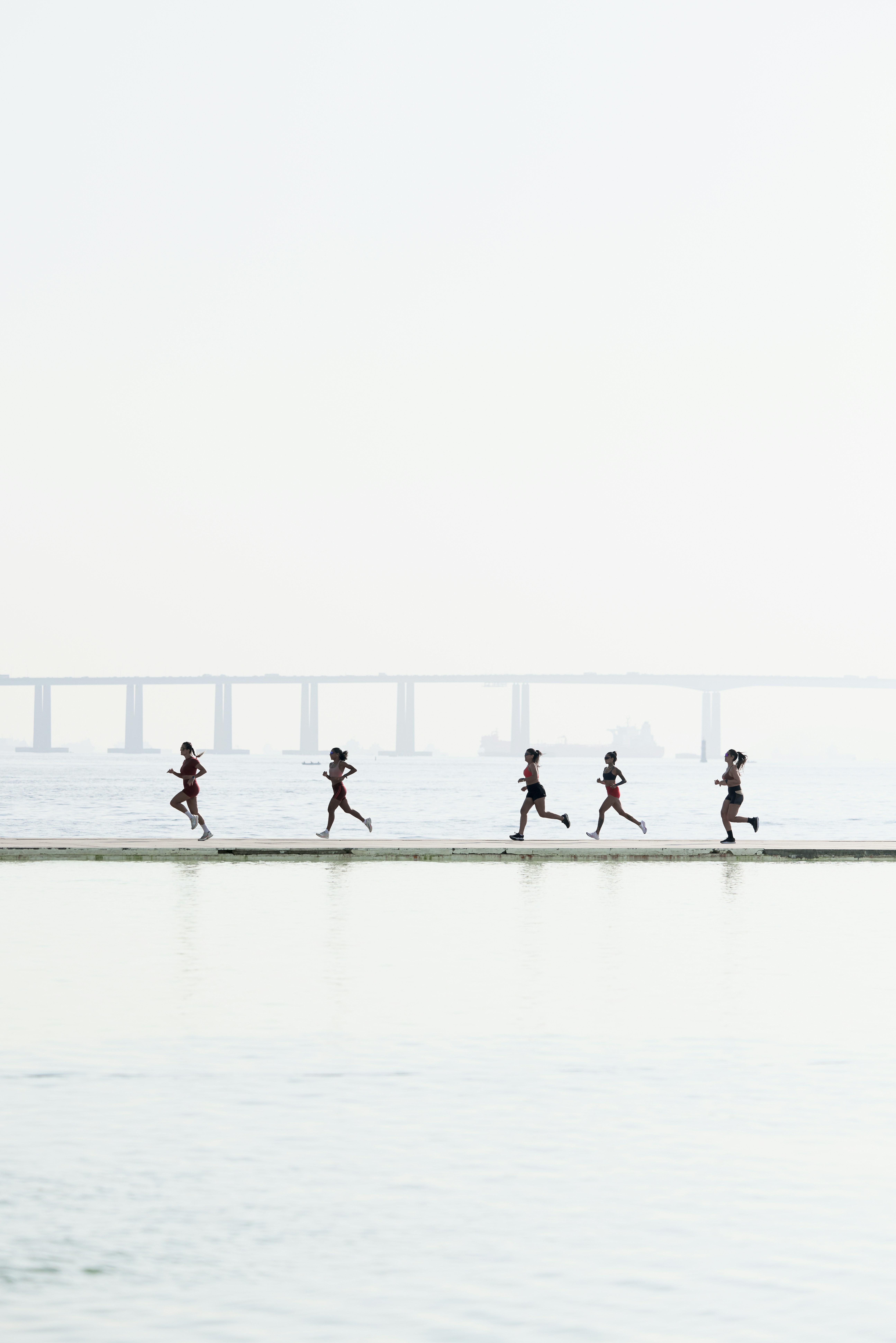 Five women jogging in a line along a narrow concrete pier over calm foggy water, wearing athletic gear in warm orange and dark tones, with a misty bridge in the hazy background