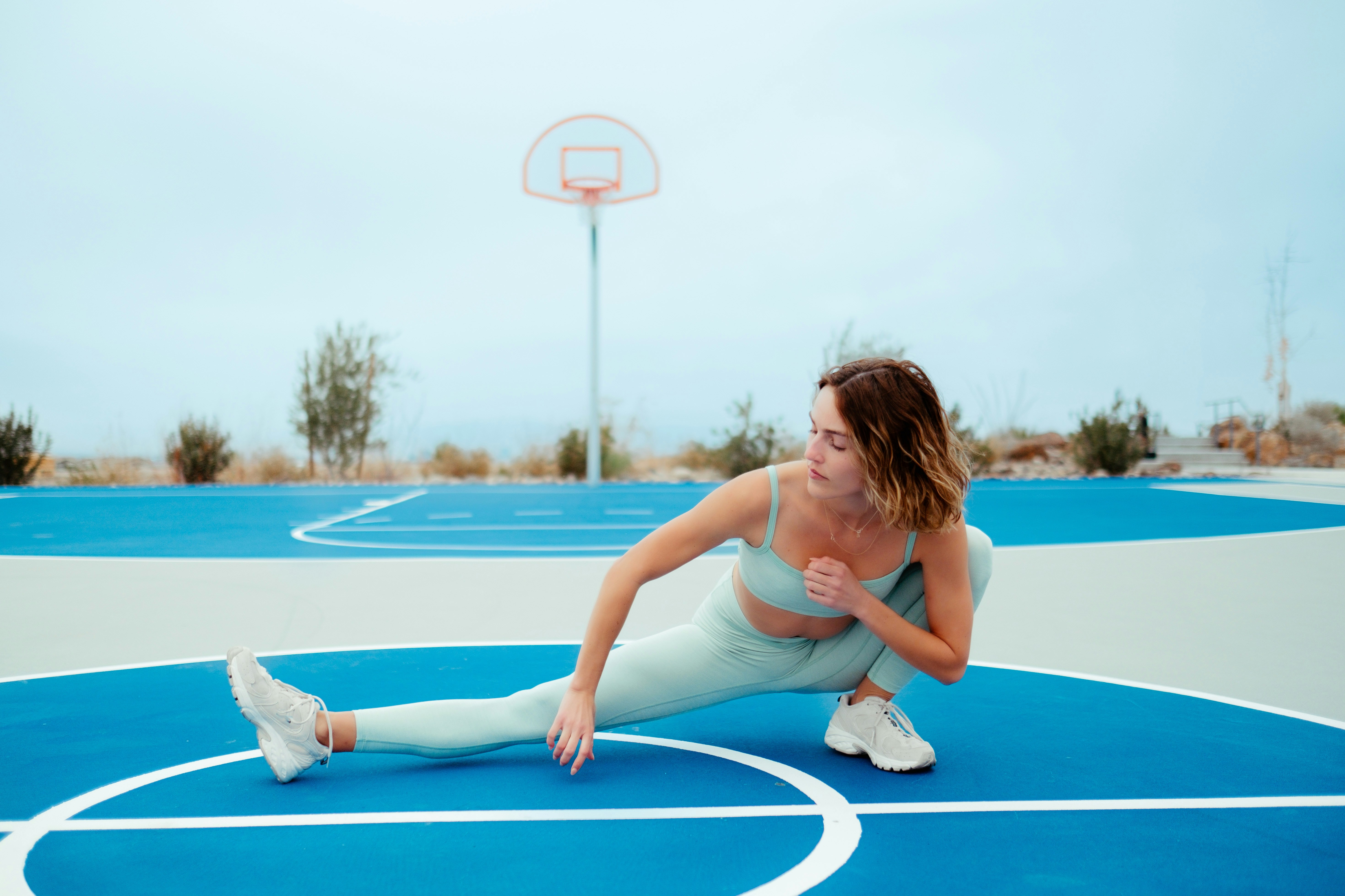 A woman in a mint-green athletic set performing a deep side lunge on a bright blue outdoor basketball court with white lines, looking downward toward her extended leg