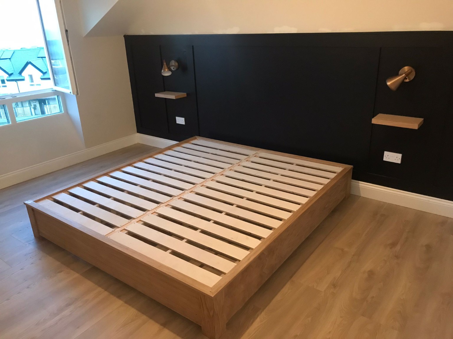 Installed oak bed frame set against a charcoal panelled headboard with floating bedside shelves and wall lights.