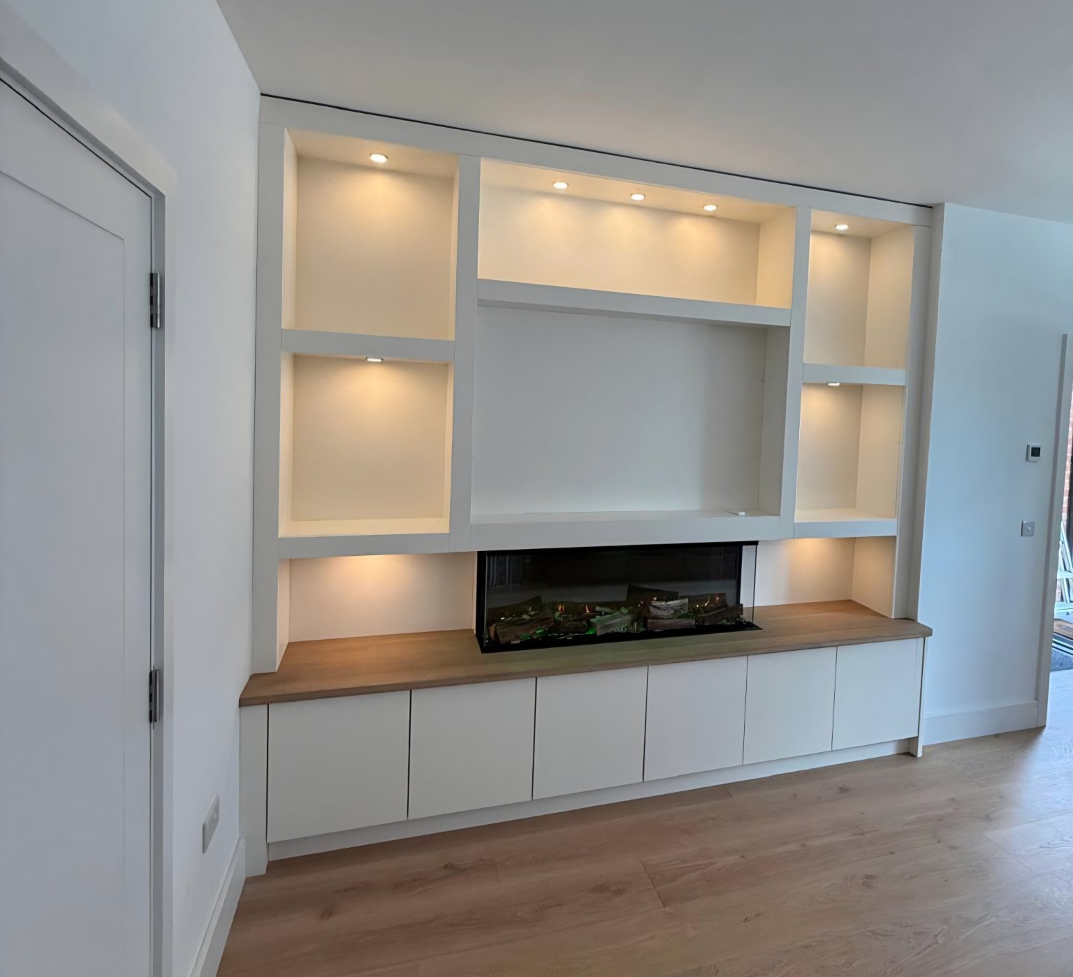 Completed off-white fitted media wall with a central fireplace, warm illuminated display shelving and an oak worktop.