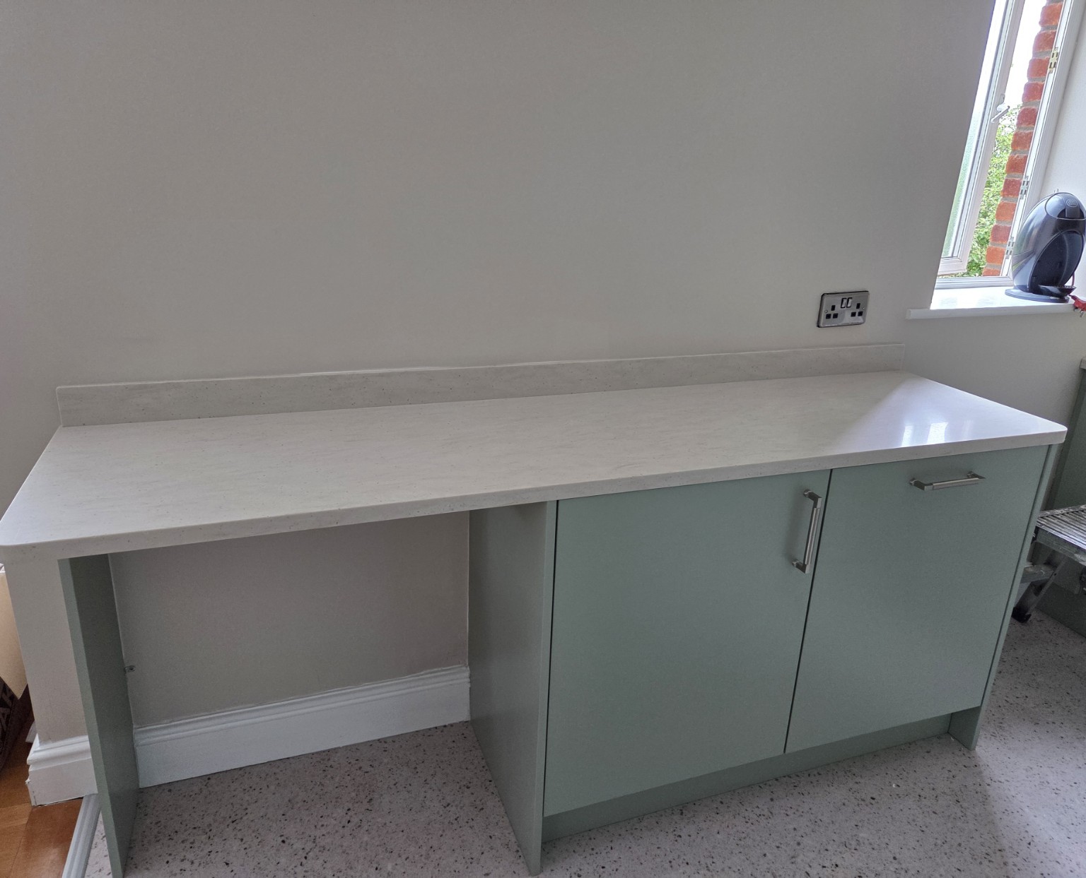 Sage-green kitchen base units with a worktop and an open space beneath part of it for accessible use.