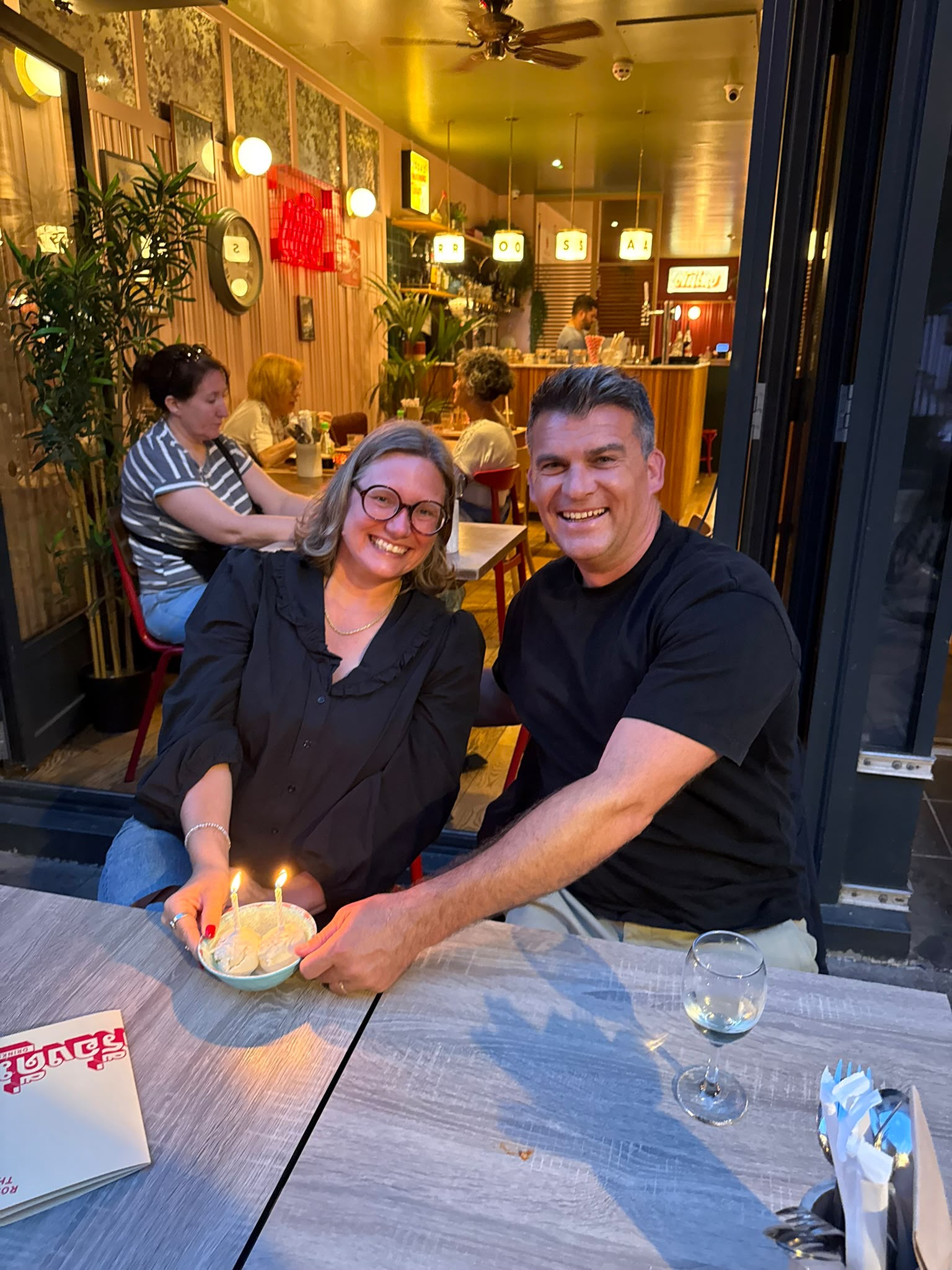 Two of the team holding a birthday dessert with lit candles at a restaurant table