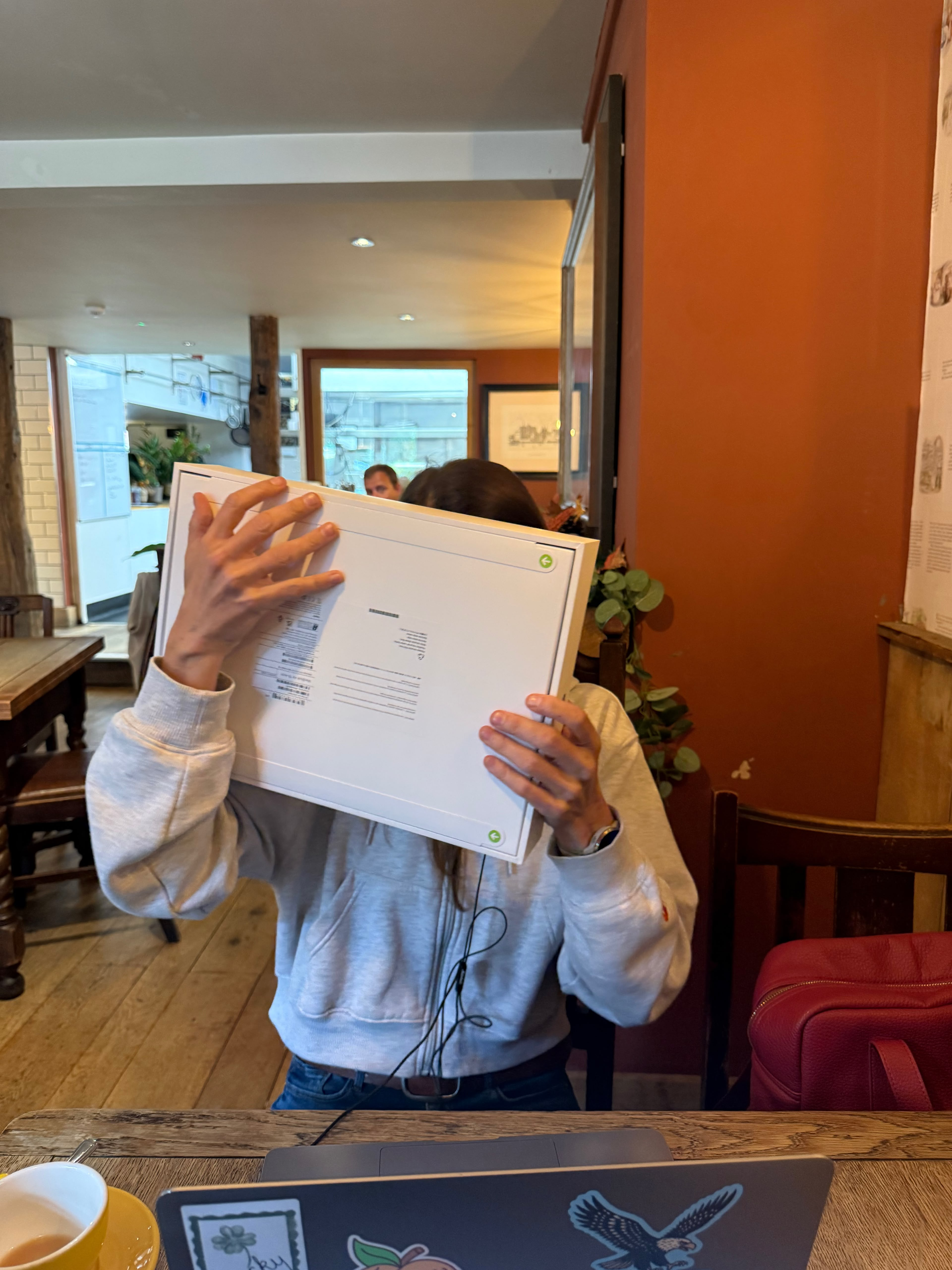 A member of the team holding a newly opened laptop box in front of their face at a cafe table