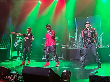 Three performers on a concert stage lit in vibrant green; the center performer sings into a microphone.