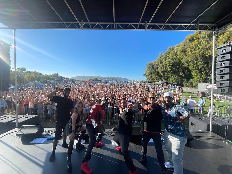 Band members posing together on an outdoor festival stage in front of a large crowd with raised hands.