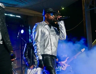 A vocalist in a metallic jacket singing into a microphone under blue stage lighting and haze, with a bassist playing alongside.