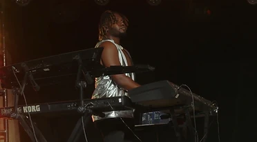 A keyboardist in a silver top playing two keyboards on a dark stage, seen in profile.