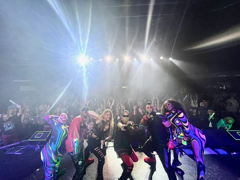 Six performers posing in a line on a dark stage before a large crowd with raised hands and glowing lights, under a bright starburst of stage lights.