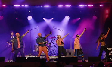 Six performers reaching upward in synchronized choreography on a concert stage under purple and magenta lighting.