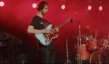 Guitarist playing a light blue electric guitar under red stage lighting, with a drummer behind the kit.