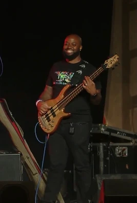 A smiling bassist playing a natural-finish bass guitar on a dark stage.