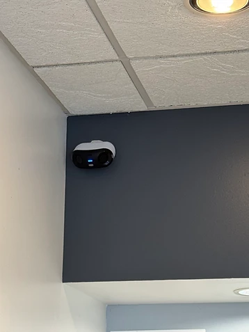 White dual-lens indoor security camera mounted on a dark gray interior corner wall