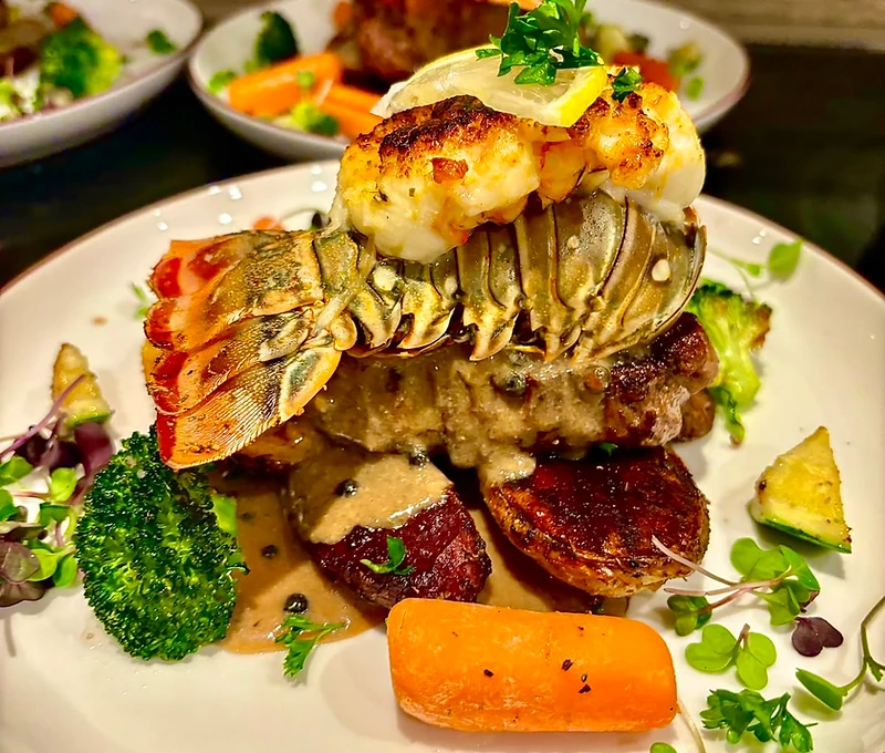 Surf and turf — seared lobster tail over steak with peppercorn sauce, roasted vegetables, and microgreens