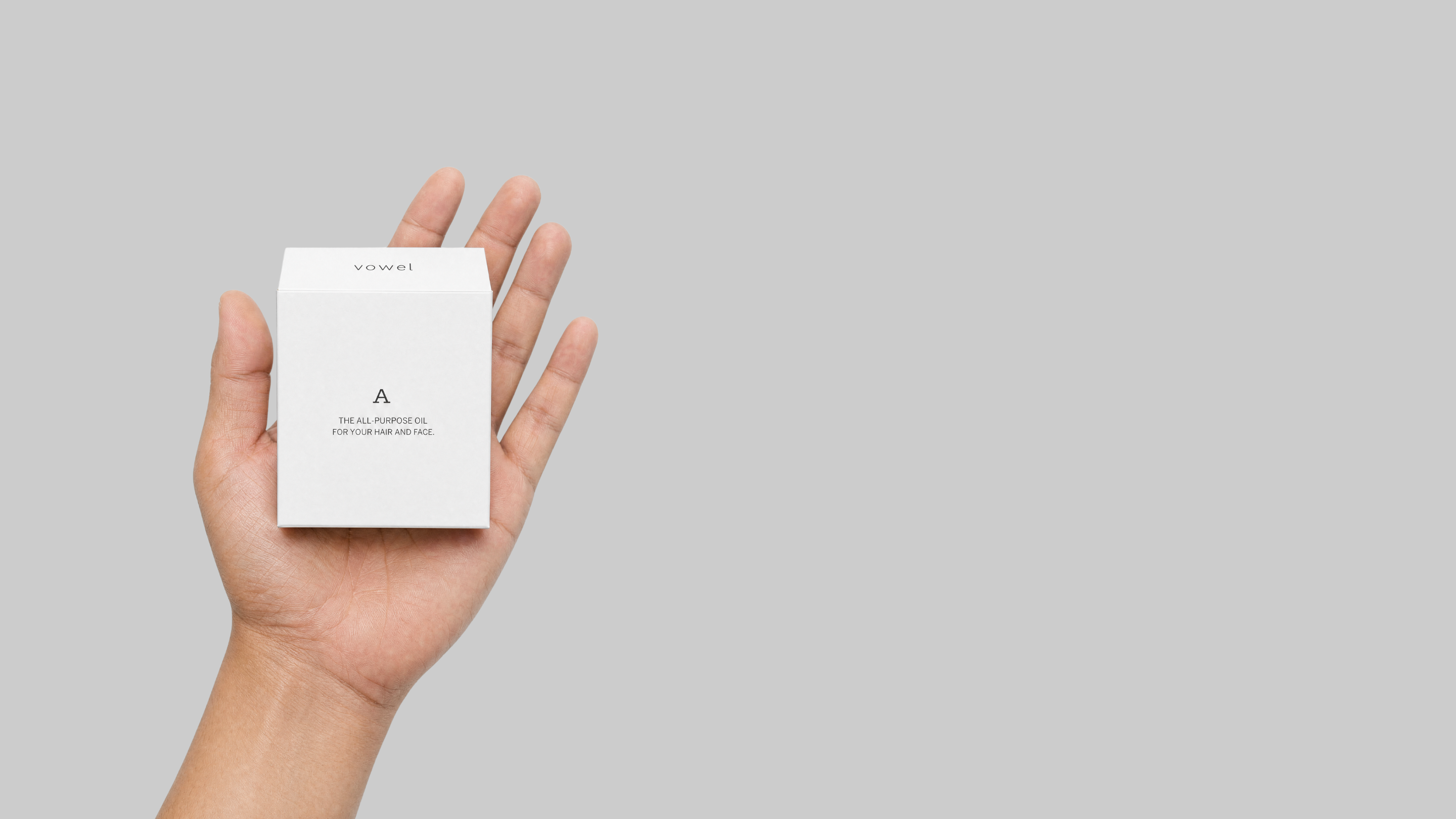 A hand holding the white A product box, the All-Purpose Oil for your hair and face, against a soft grey background