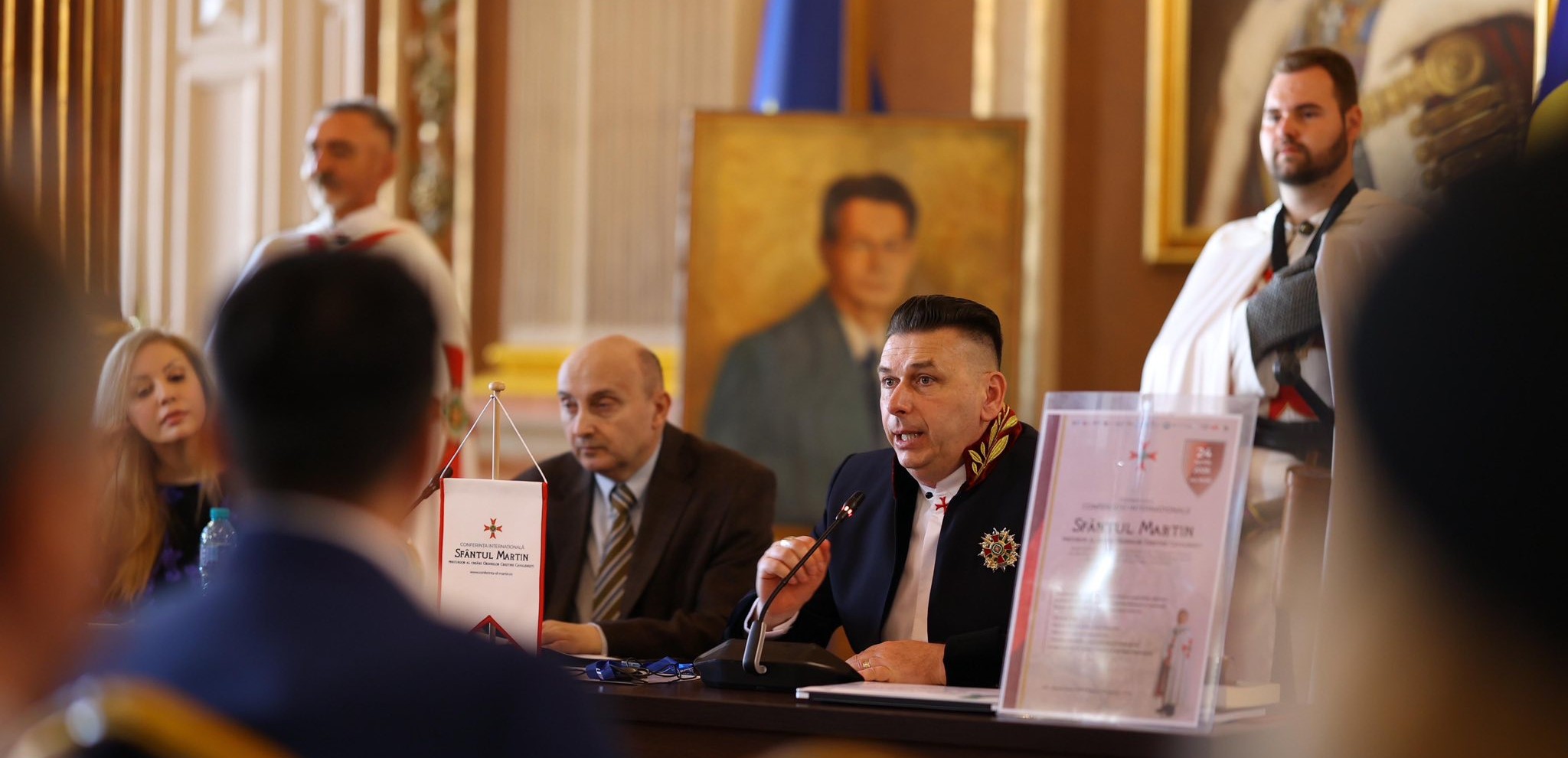 Helena B. Scott's conference presentation at the Arad conference, Romania — a formal Templar-guarded conference setting with the Sfântul Martin event poster visible.