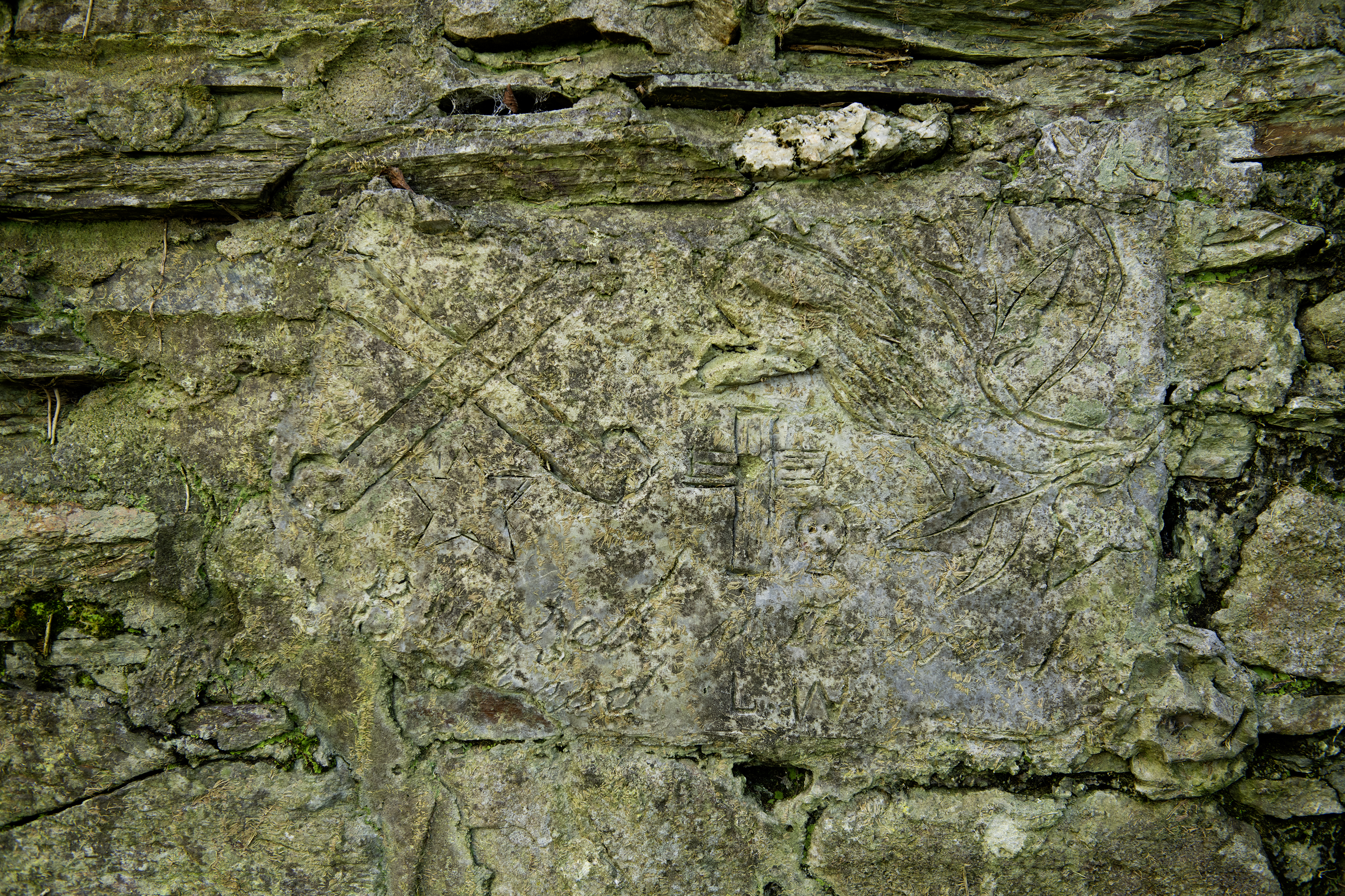 A lichen-covered stone slab set in a dry-stone wall, incised with a cross pattée, a crossed staff emblem, a foliage motif and the faint initials L.W.