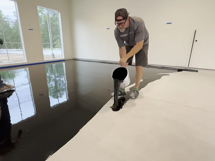 Installer pouring a black moisture vapor barrier coating onto a primed concrete floor while a second installer rolls it out
